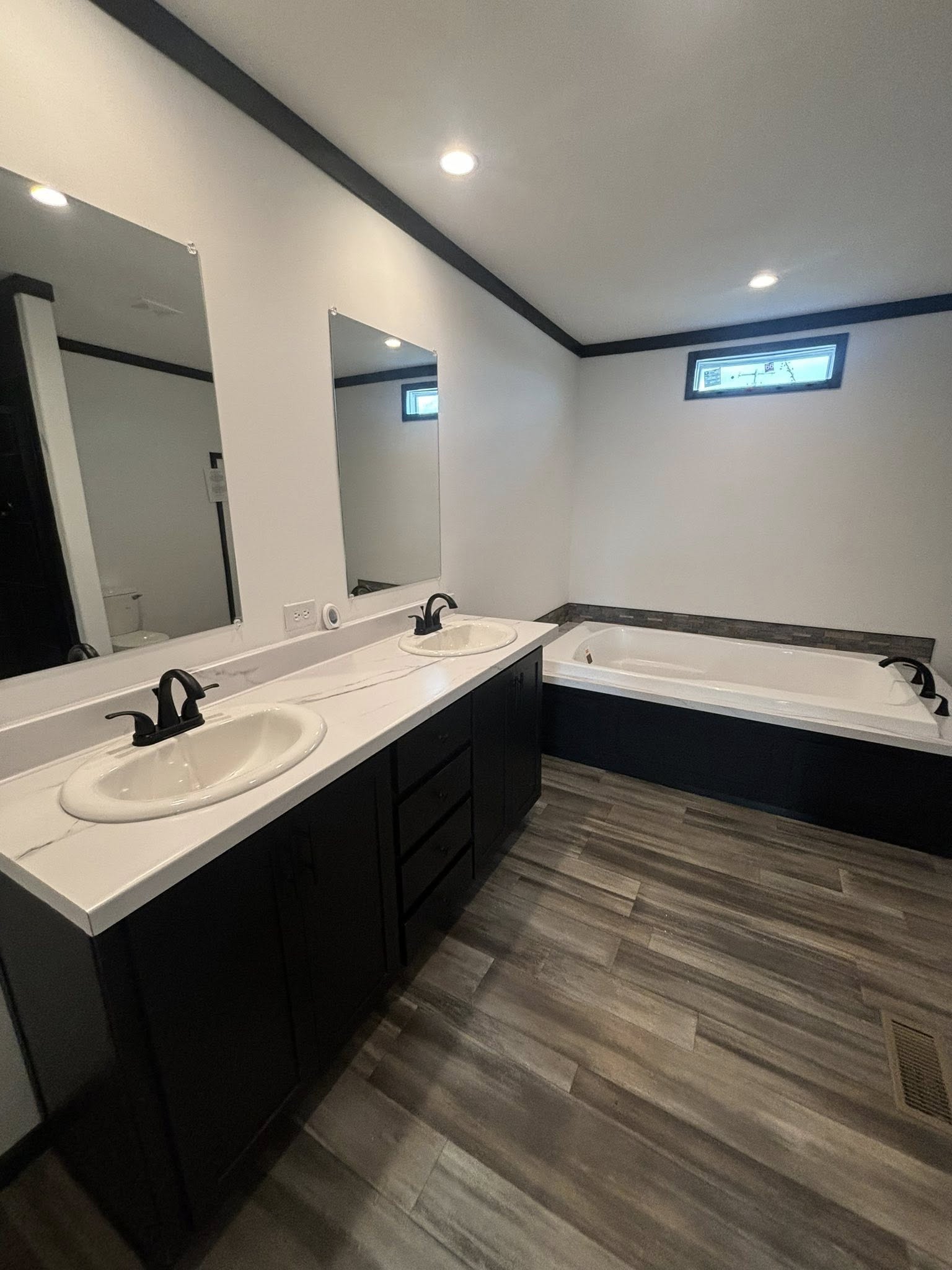 Modern bathroom with a sleek double vanity and dark cabinetry. Two large mirrors above sinks enhance space. A sunken bathtub is on the right, creating a serene ambiance.