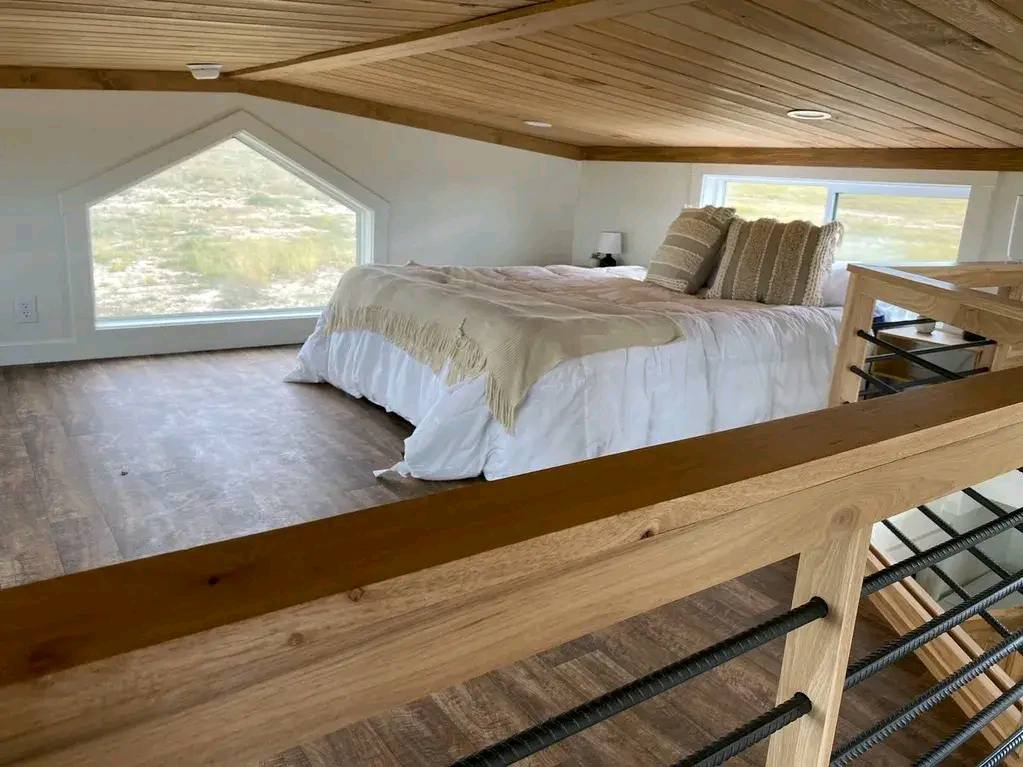 A cozy loft bedroom with a sloped wooden ceiling and large windows. It features a neatly made bed with beige pillows and a throw, exuding a warm, serene vibe.
