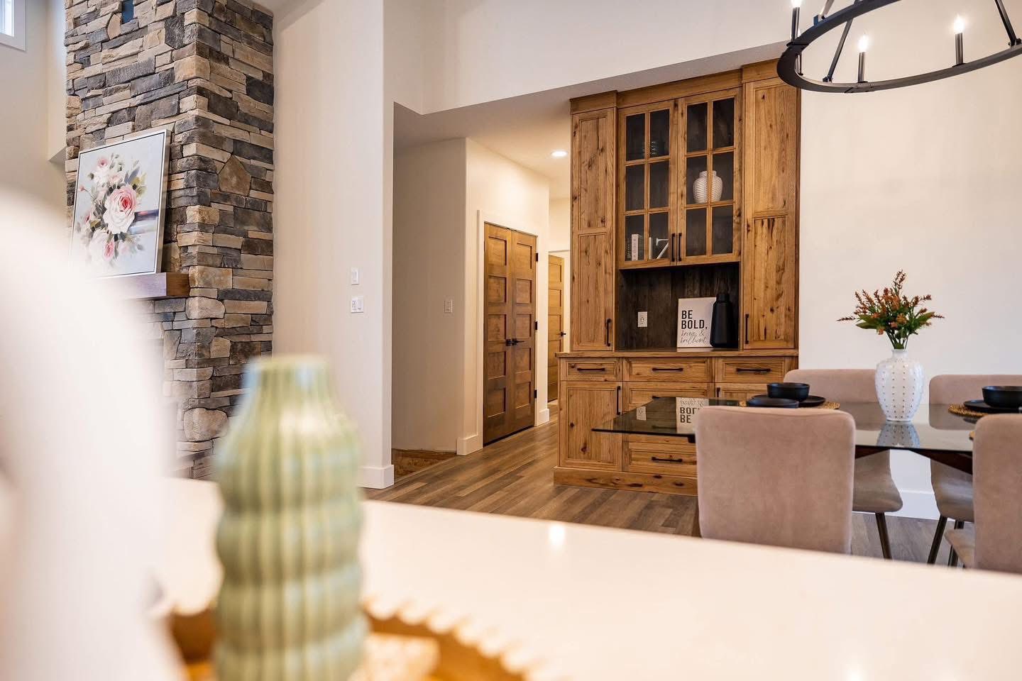 Warm, cozy dining area with wood accents. Features a stone fireplace, rustic cabinet, floral decor, and a table with beige chairs, evoking a welcoming atmosphere.