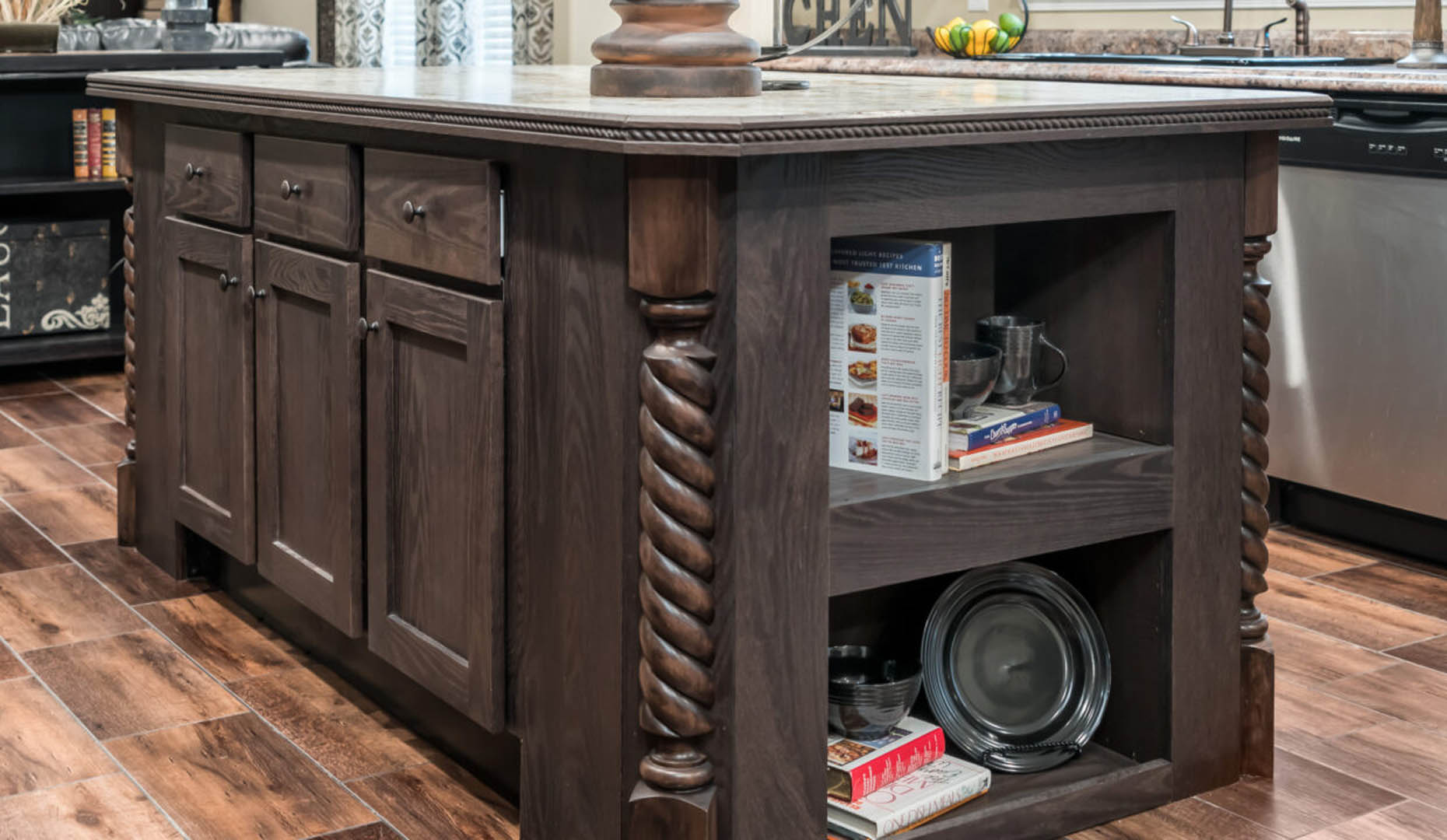 A rustic wooden kitchen island with twisted column legs, cabinetry, and open shelving. The shelves hold cookbooks and dishes. The tone feels warm and inviting.
