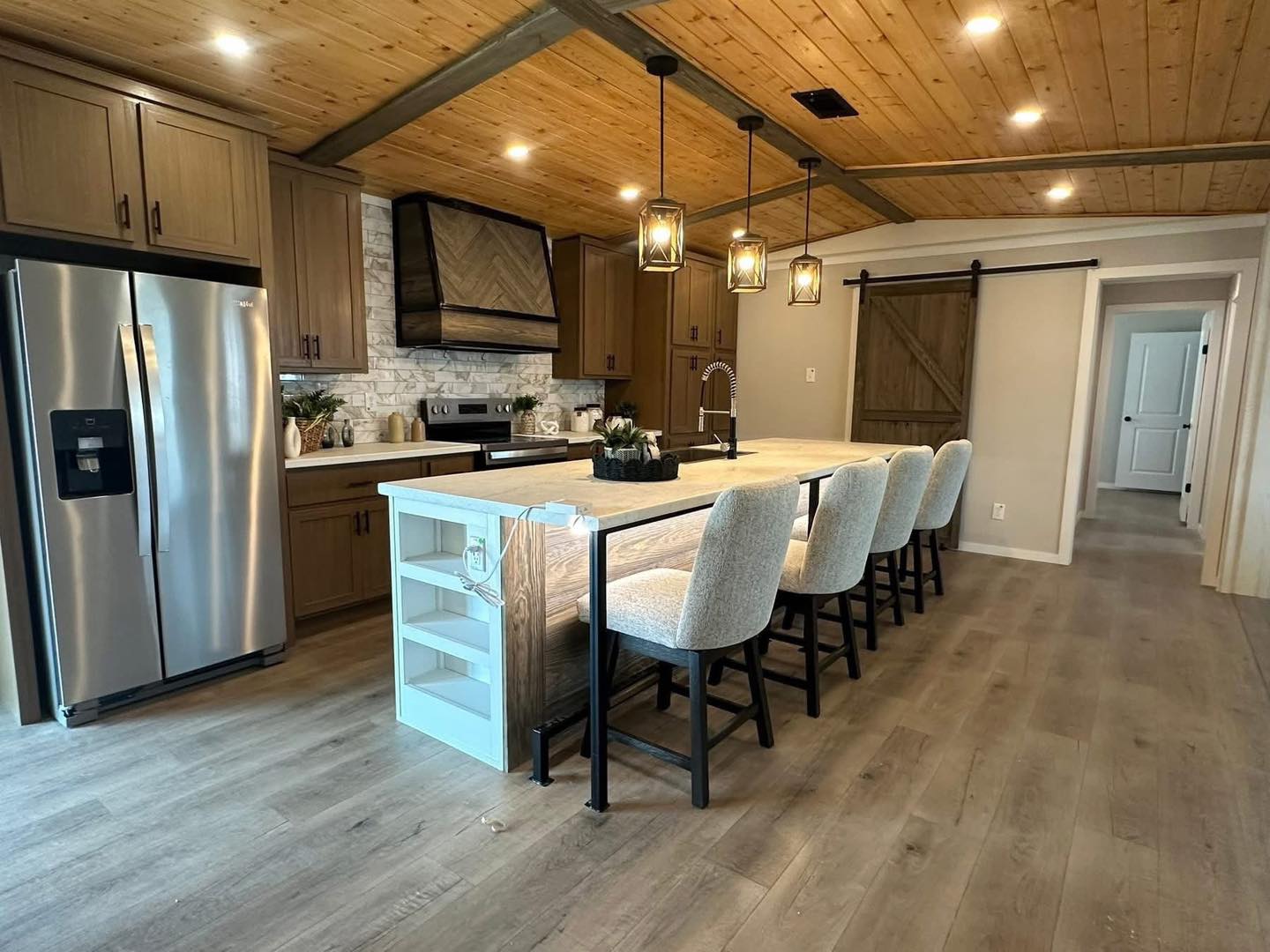 Modern kitchen with wooden ceiling and floors, featuring a large island with four cushioned stools, pendant lights, stainless steel appliances, and a rustic sliding barn door. Cozy and inviting atmosphere.