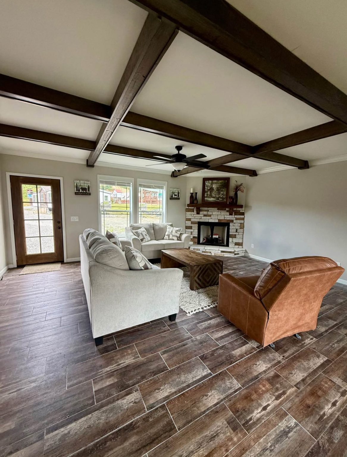 Spacious living room with wood beam ceiling, light gray sofas, brown armchair, and a stone fireplace. Bright light from large windows. Cozy and inviting.