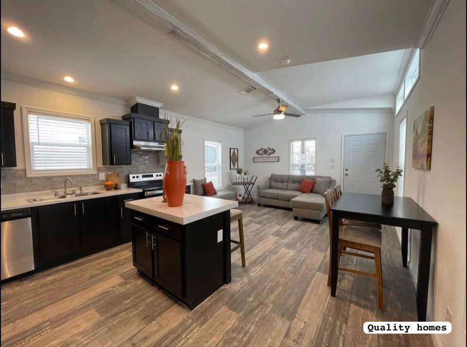 Modern open-plan kitchen and living room with wood flooring. Black cabinets, central island with a plant, gray sofa, and wall art create a cozy, stylish atmosphere.
