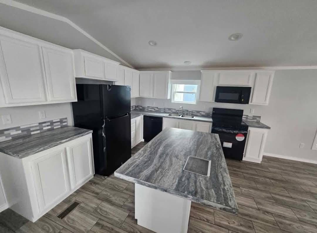 Modern kitchen with white cabinets, gray countertops, and wood-patterned flooring. Features a central island, black appliances, and a bright window.