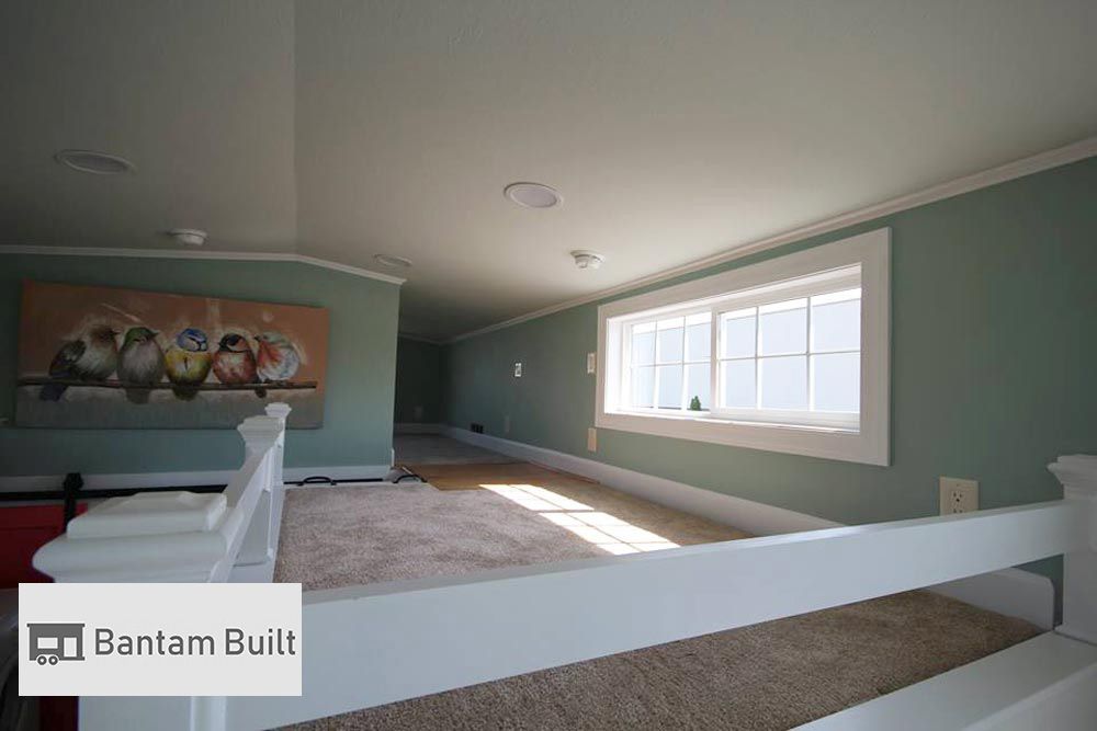 A cozy, carpeted loft space with teal walls and a low ceiling. A rectangle window casts sunlight inside, while a bird painting adds charm.