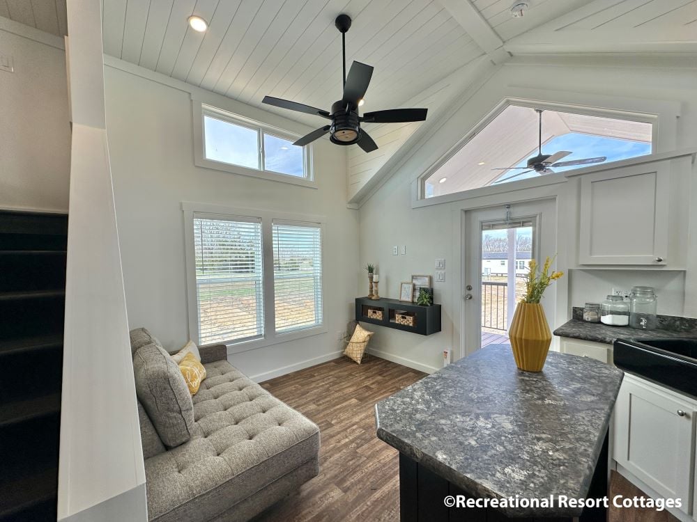 Inviting tiny home interior with high ceilings and ample windows. Features a cozy gray sectional, modern kitchen with black counters, and bright decor.