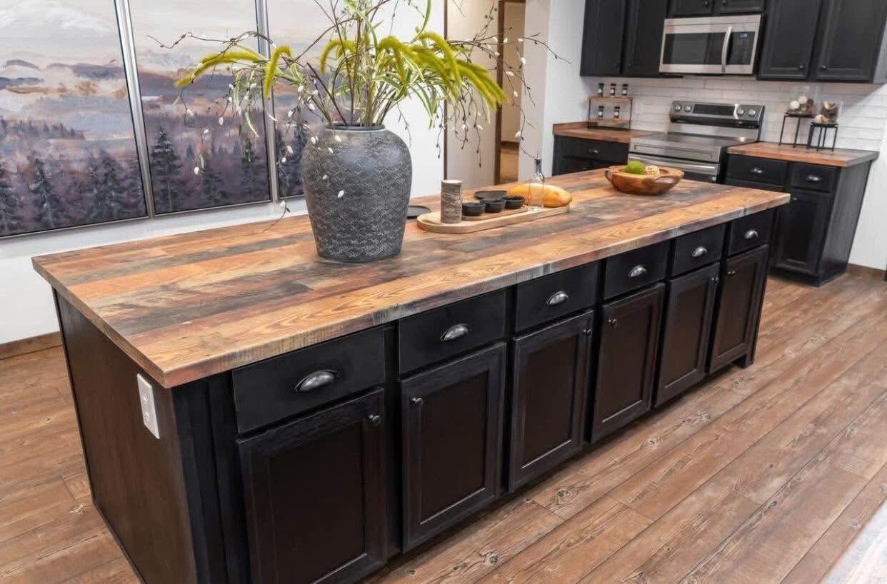Rustic kitchen with a wood island featuring dark cabinets. A large vase with green plants sits on the counter. The background shows modern appliances.