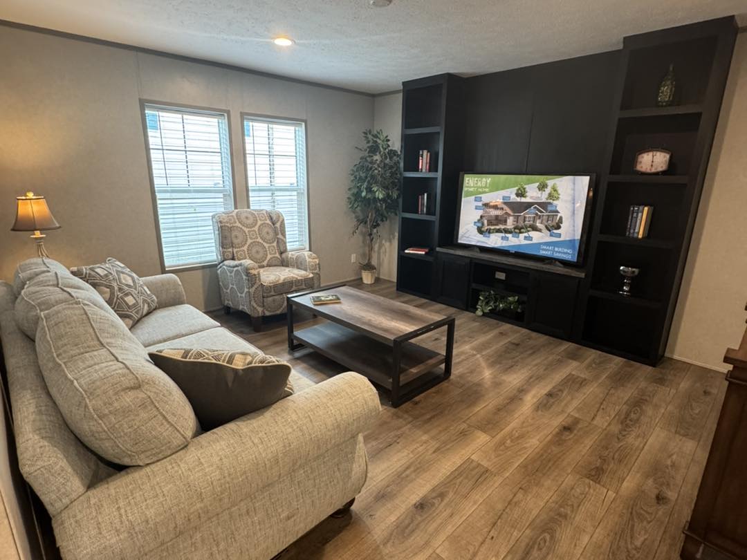 Cozy living room with a beige sofa and patterned armchair. A TV displays a home image on a dark entertainment center. Warm lighting and wood floors.