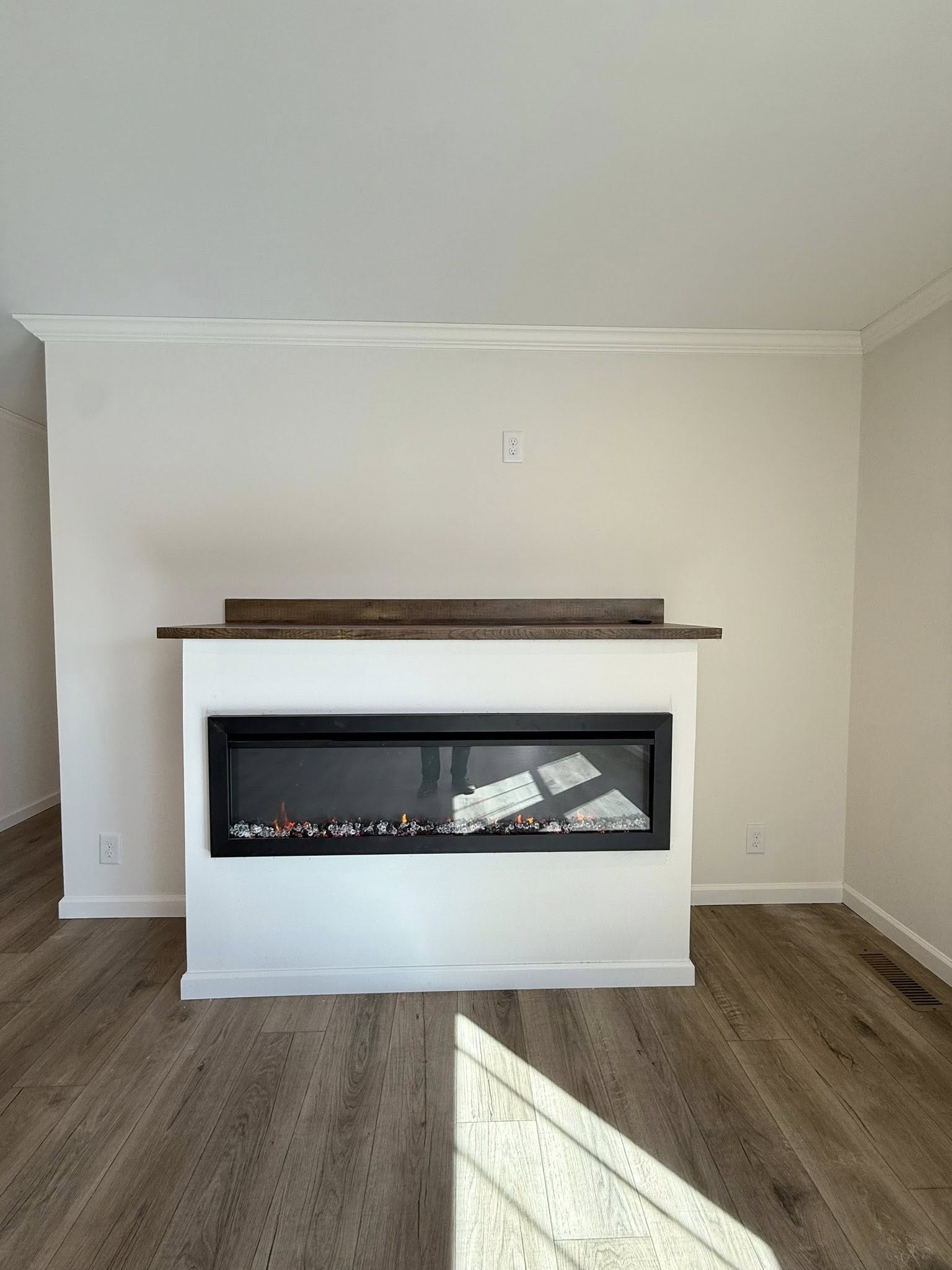 Modern living room with a sleek electric fireplace inset into a white wall, beneath a wooden mantel. Sunlight casts a shadow on the light wood floor.