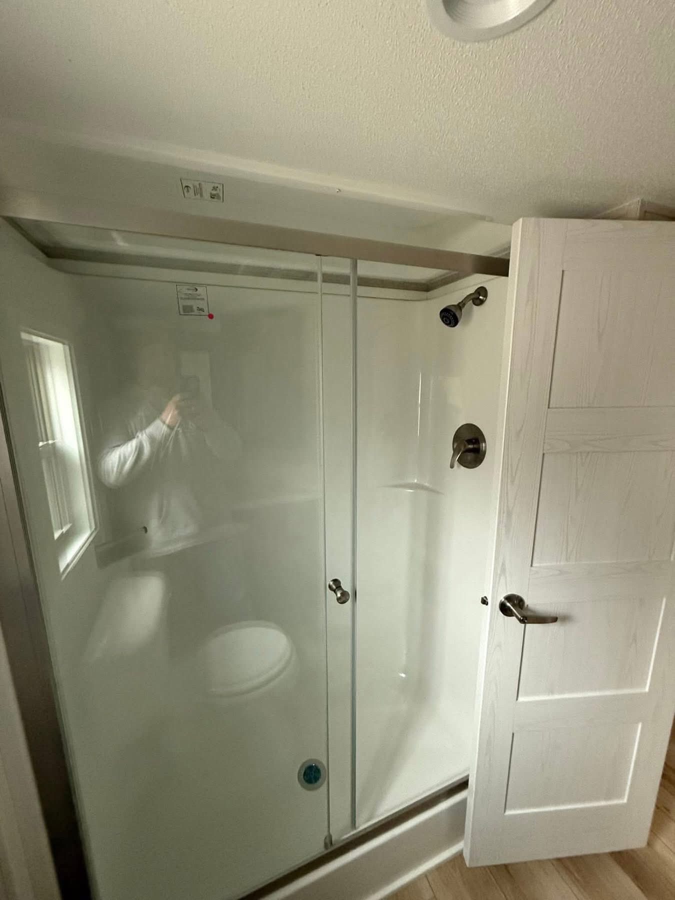 Small white shower stall with sliding glass door, silver fixtures, and an open white wooden door. A dimly lit room with a clean, minimalist feel.