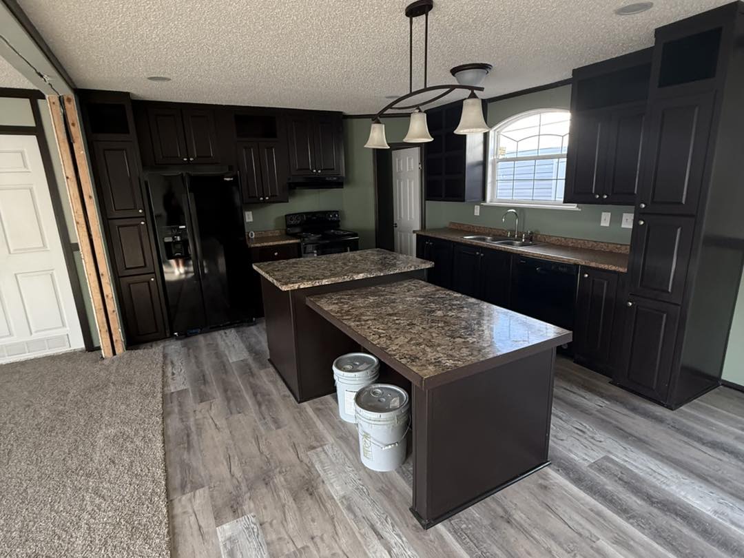 Modern kitchen with dark cabinets, granite countertops, and an island with pendant lights. Light wood flooring contrasts with dark tones.
