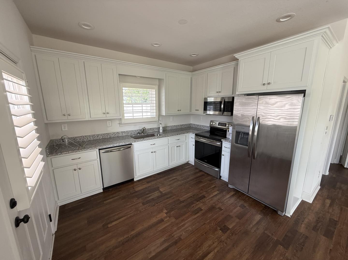 Modern kitchen with white cabinets, stainless steel appliances, and granite countertops, featuring a hardwood floor and a window with blinds.
