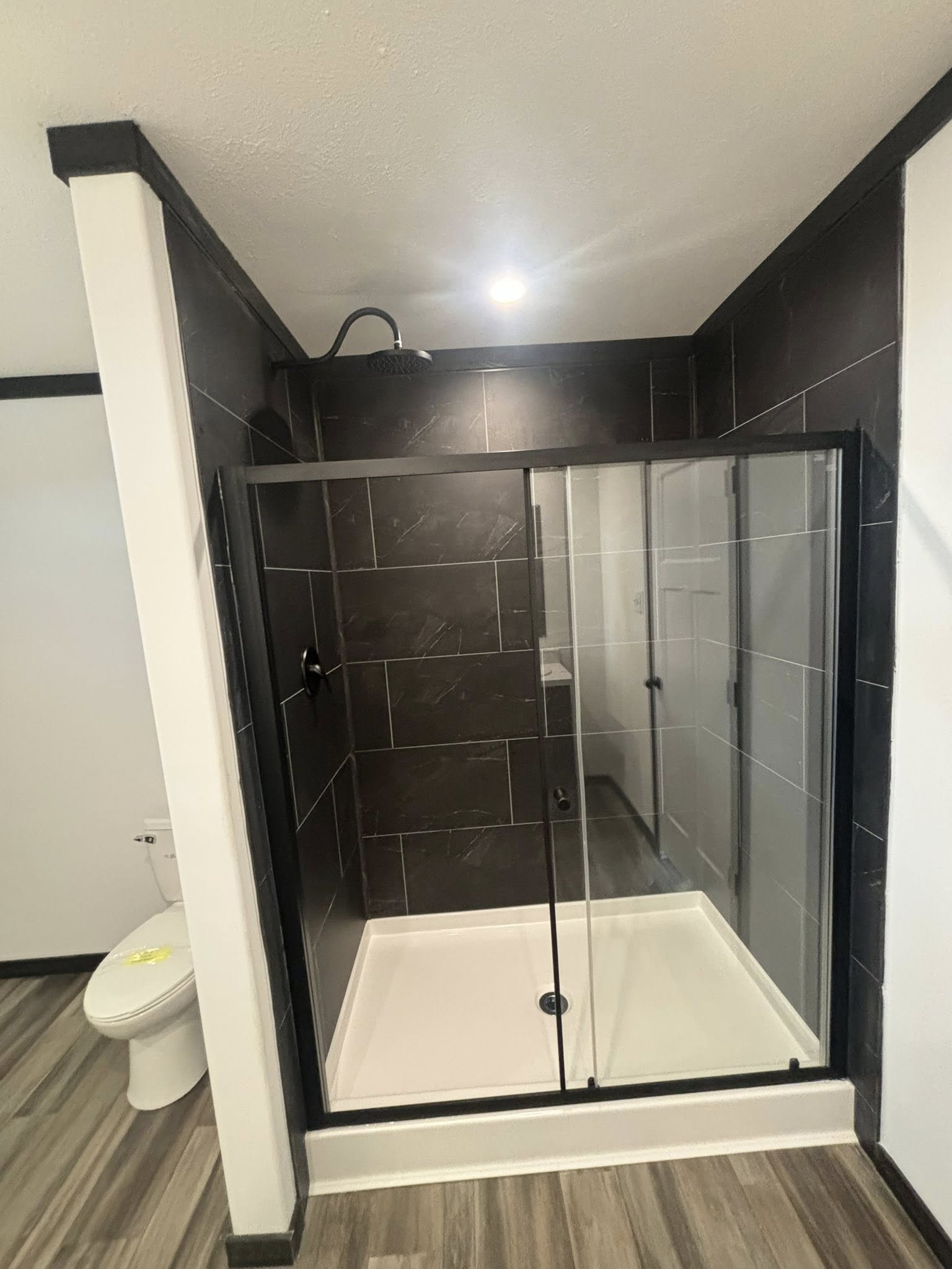 Modern bathroom with glass shower enclosure. Black tiled walls contrast with white shower base. Adjacent is a toilet and wood-patterned floor. Bright lighting.
