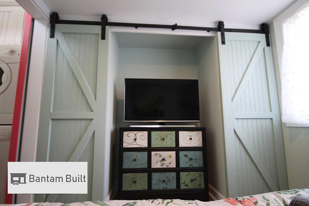 Alt text: A cozy space features a flat-screen TV on a black dresser with floral patterns. Light green sliding barn doors frame the nook, creating a rustic feel.