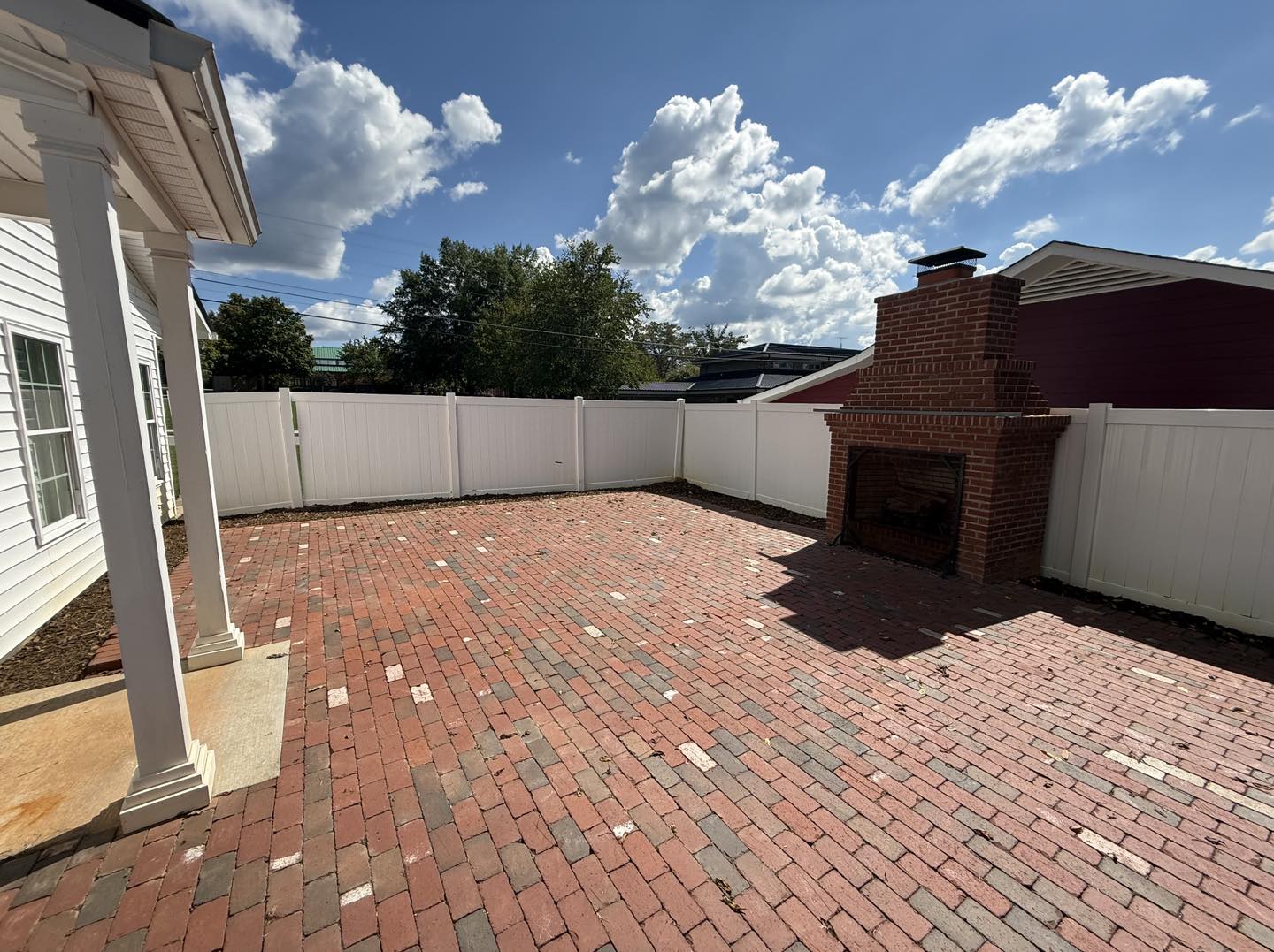 Sunny brick patio with a red brick outdoor fireplace and white privacy fence. Blue sky with puffy clouds adds a calm, open-air feel.