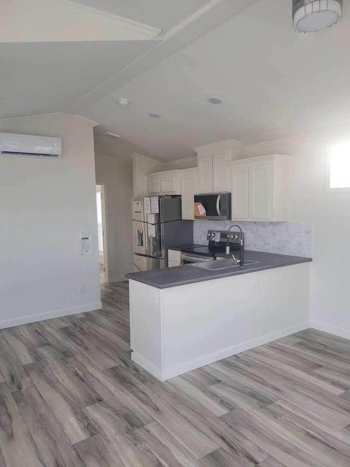 Modern kitchen with white cabinets, gray countertops, and stainless steel appliances. Light wood flooring and a bright window enhance the airy ambiance.