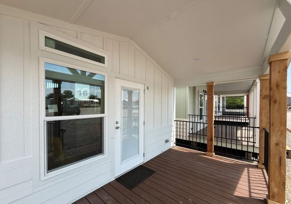 Covered porch with wooden deck and light siding, featuring a glass door and window. Wooden posts support the roof. Bright, welcoming atmosphere.