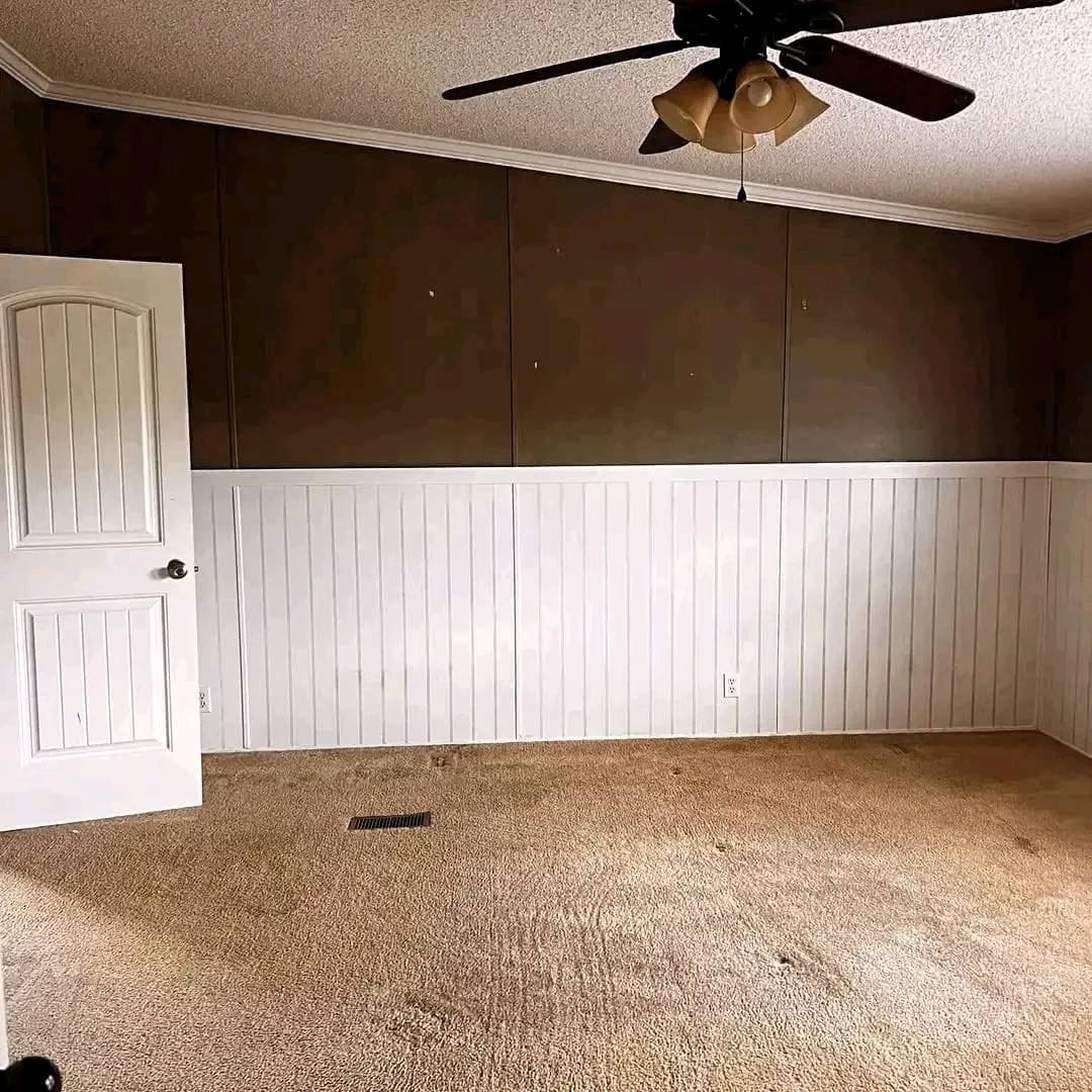 Empty room with beige carpet, brown upper walls, and white vertical paneling. An open door and ceiling fan create a neutral, quiet atmosphere.