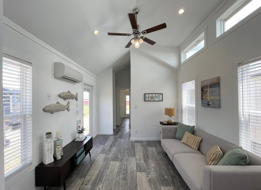 Bright, cozy living room with a vaulted ceiling and wood flooring. A gray sofa with cushions sits near a window. Nautical decor and a "Home" sign adorn white walls.