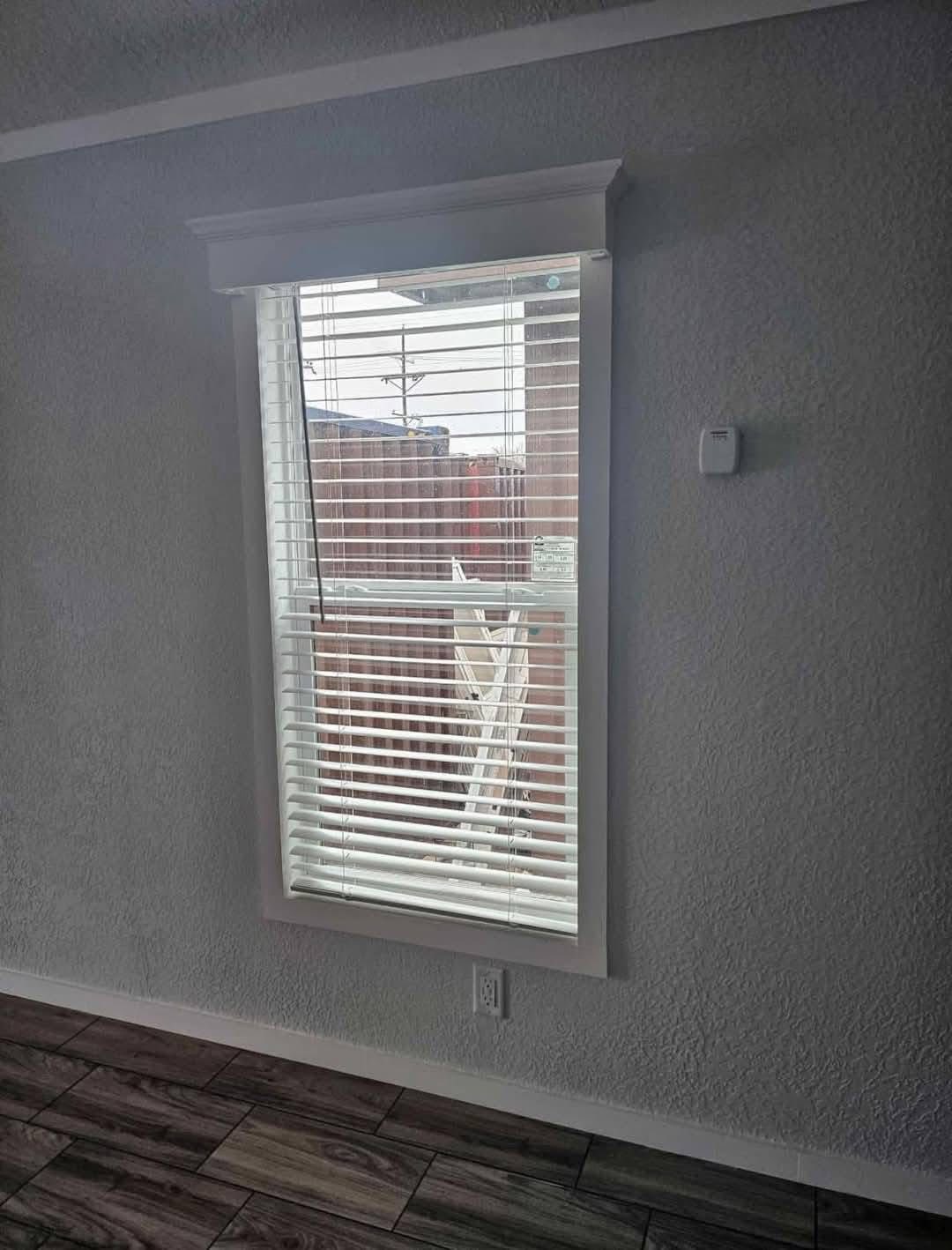 A window with white blinds in a room with textured gray walls and wood flooring. Light filters through, casting shadows and a ladder is visible outside.