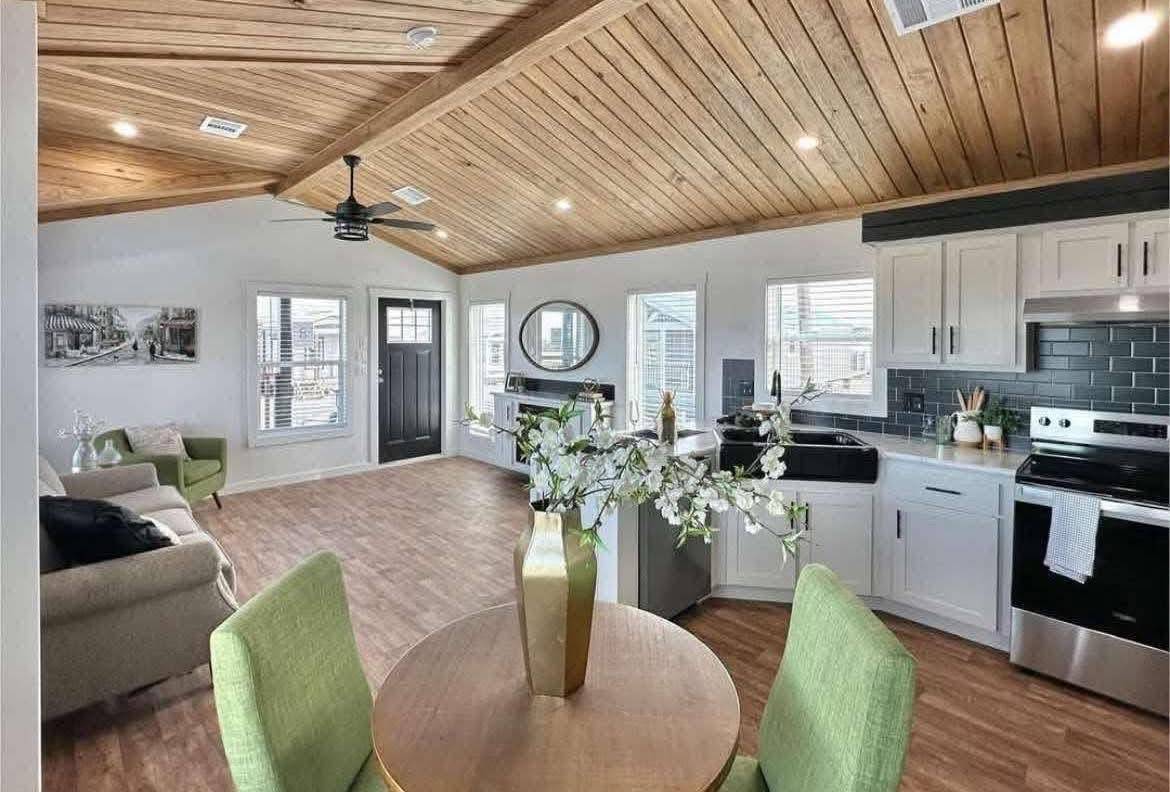 Modern open-concept living space with wooden vaulted ceiling. Features a kitchen, round table with flowers, and cozy seating area. Bright, airy ambiance.