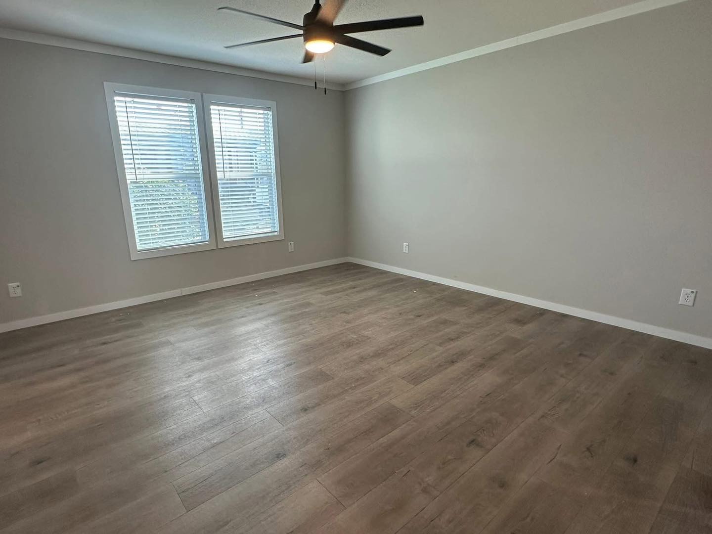 An empty room with light gray walls, wooden floor, and a ceiling fan. Two windows with blinds allow soft natural light, creating a calm atmosphere.