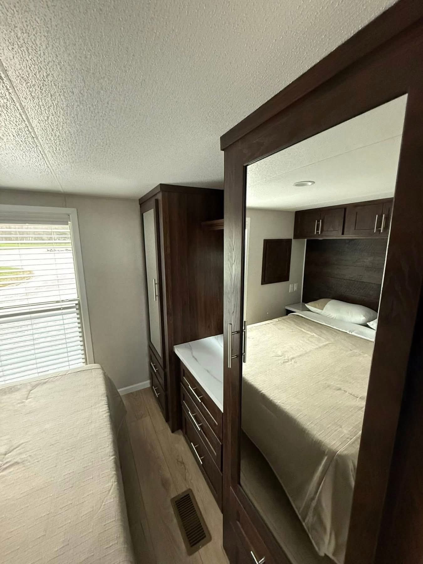 Compact bedroom with mirrored wardrobe reflecting a bed with beige linens. Dark wood cabinets and natural light from a window create a cozy atmosphere.