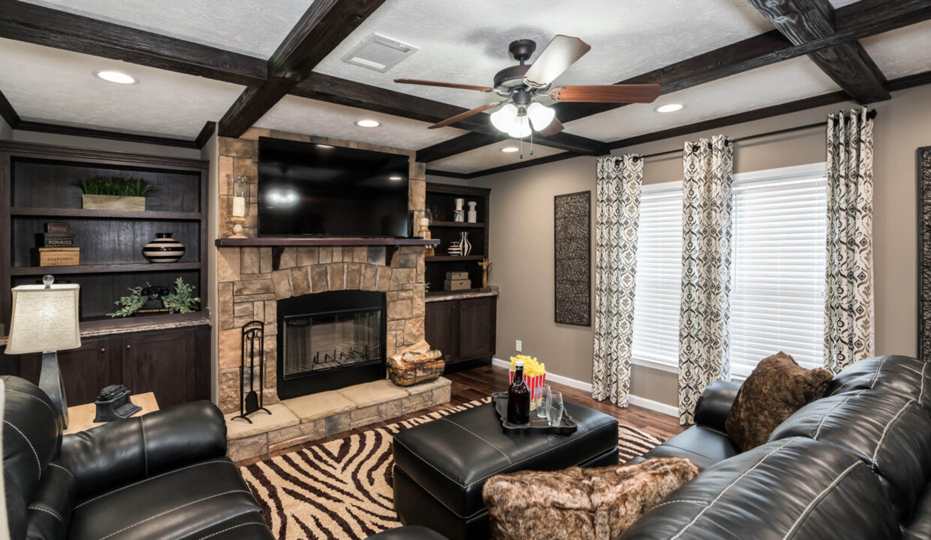 A cozy living room features a stone fireplace, black leather sofas, and a zebra-patterned rug. Large windows with patterned curtains allow natural light to fill the room.