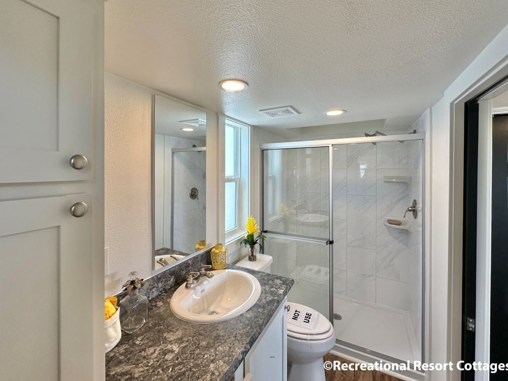 Modern bathroom with a granite countertop, white sink, and a glass-door shower. Bright lighting, yellow flower accents, and a sleek, clean design convey freshness.