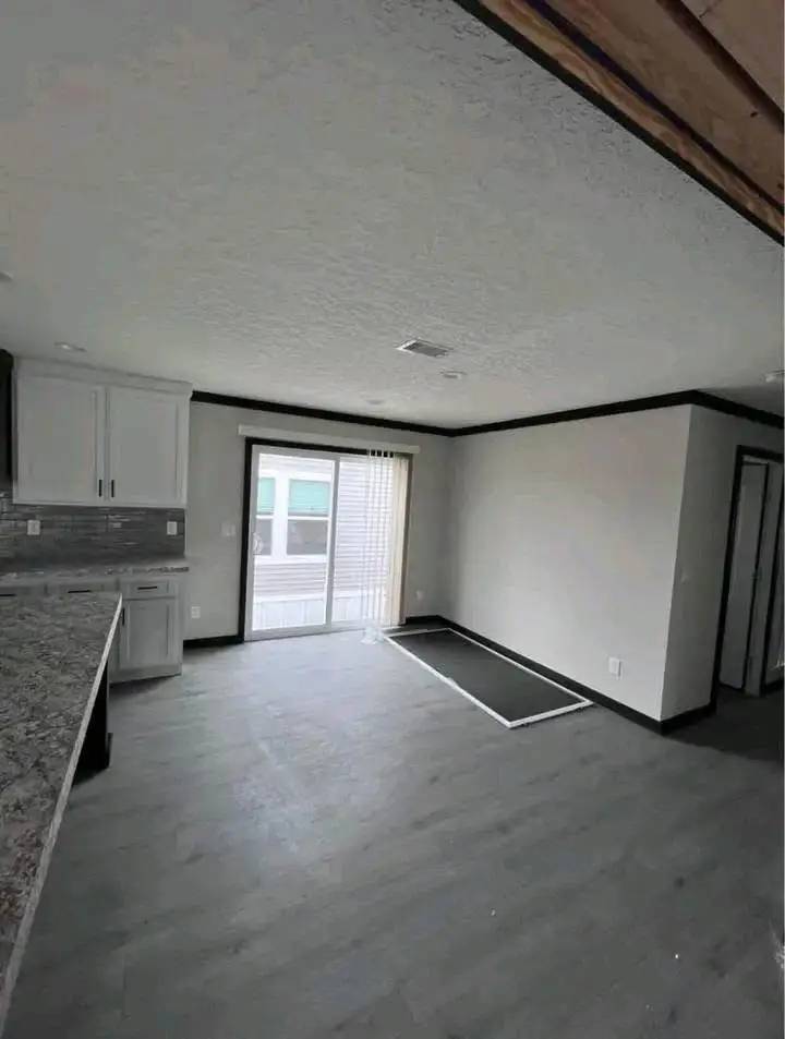 Modern, unfurnished room with dark wood floor and light gray walls. Kitchen cabinets on left, sliding glass doors back, screen on floor, minimalist feel.