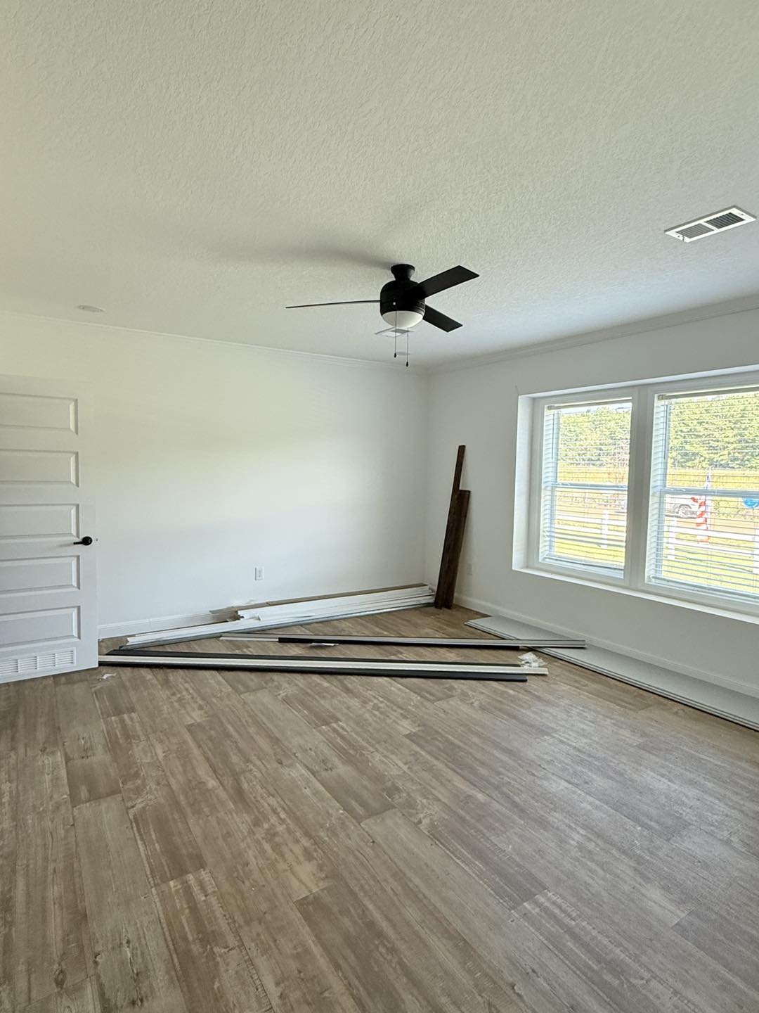 Empty room with light wood flooring, white walls, and a black ceiling fan. Large windows let in natural light. Flooring planks are on the floor.