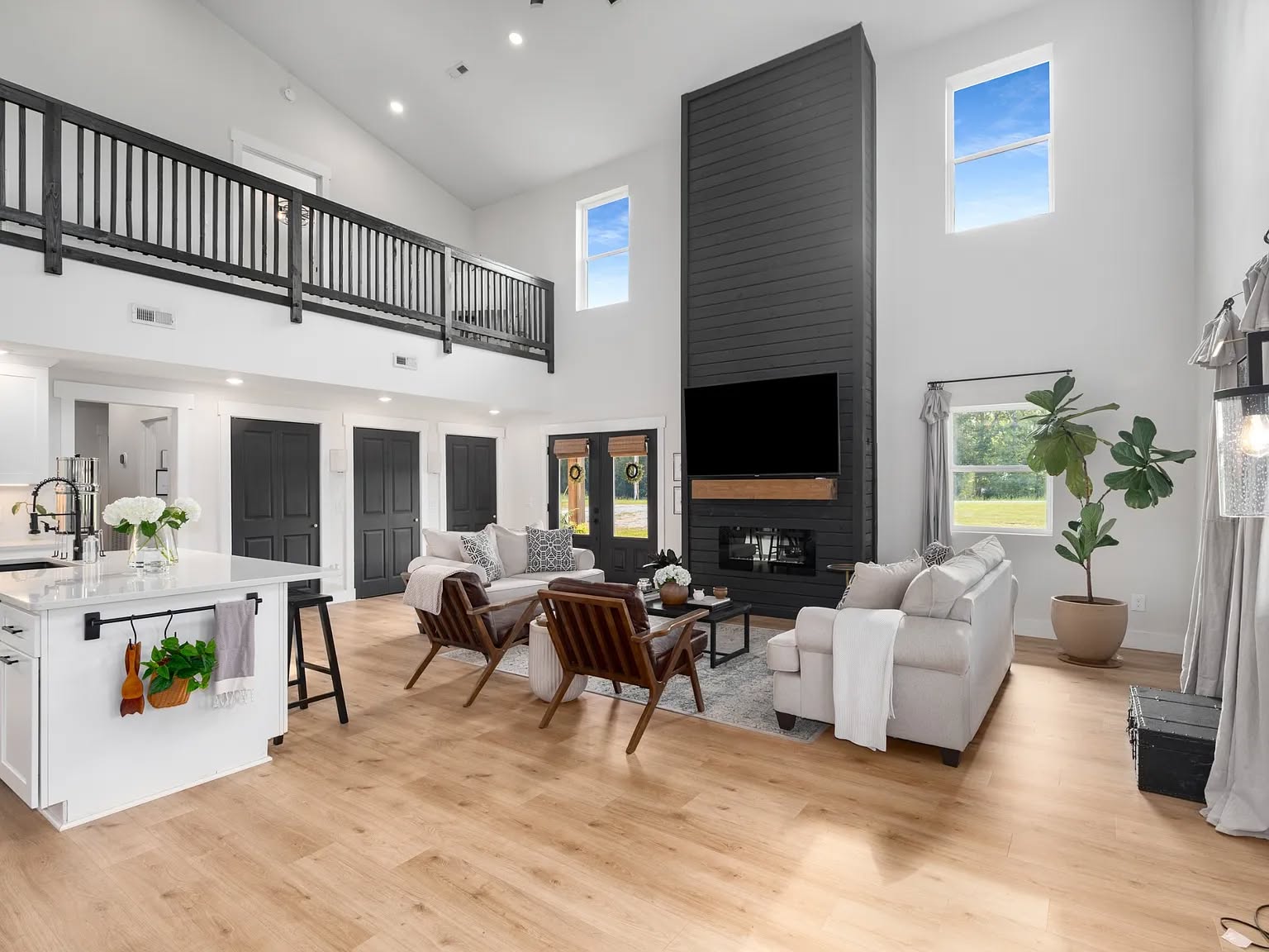 Spacious modern living room with high ceilings features a black accent wall with a large TV and fireplace. Light wood floors, white walls, and cozy furniture create an inviting atmosphere. Two-story windows and balcony enhance the airy feel.
