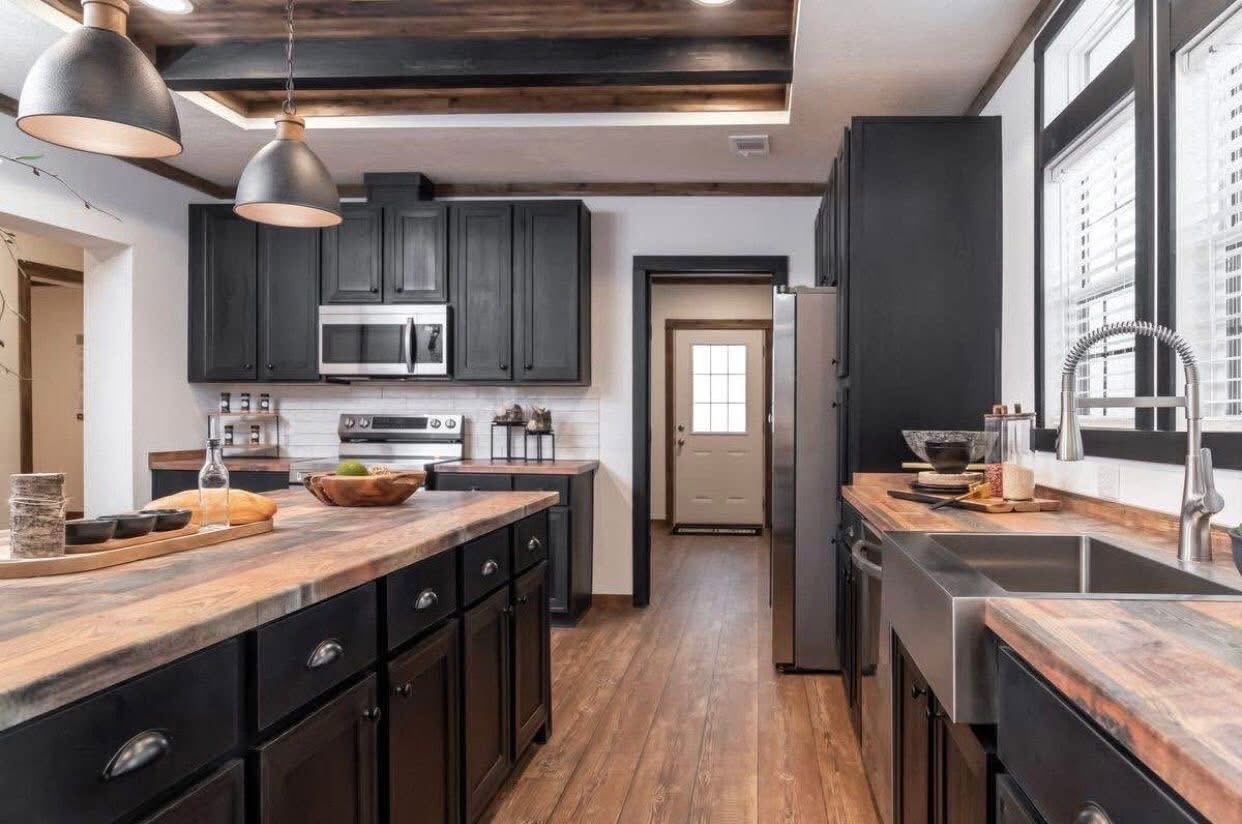 Modern kitchen with dark cabinets, wood countertops, and island under pendant lights. Stainless steel appliances and large sink by the window. Warm and inviting.