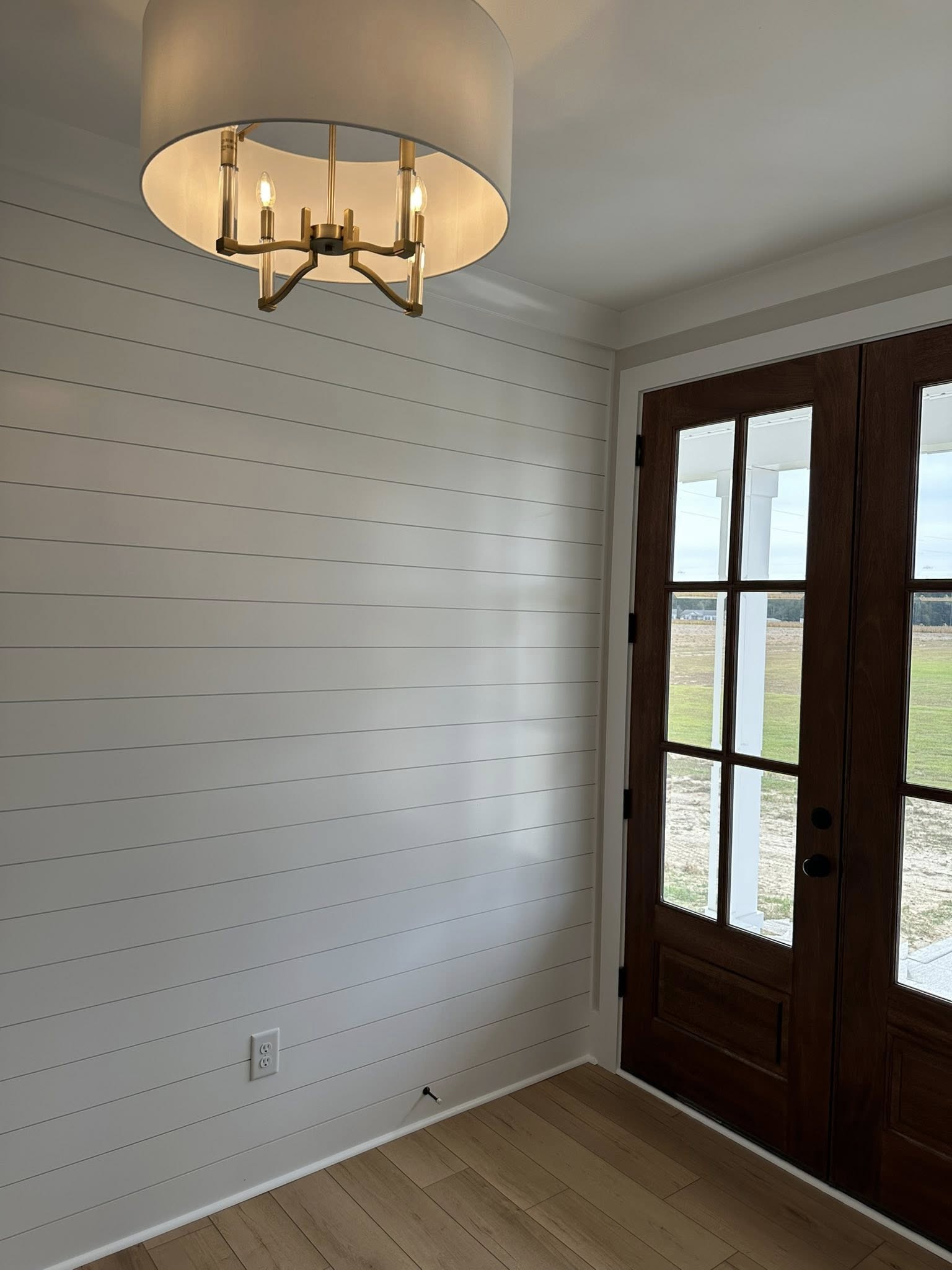 Room with a modern chandelier, white shiplap walls, and a wooden French door. Outside view shows a field under a cloudy sky, evoking calm.