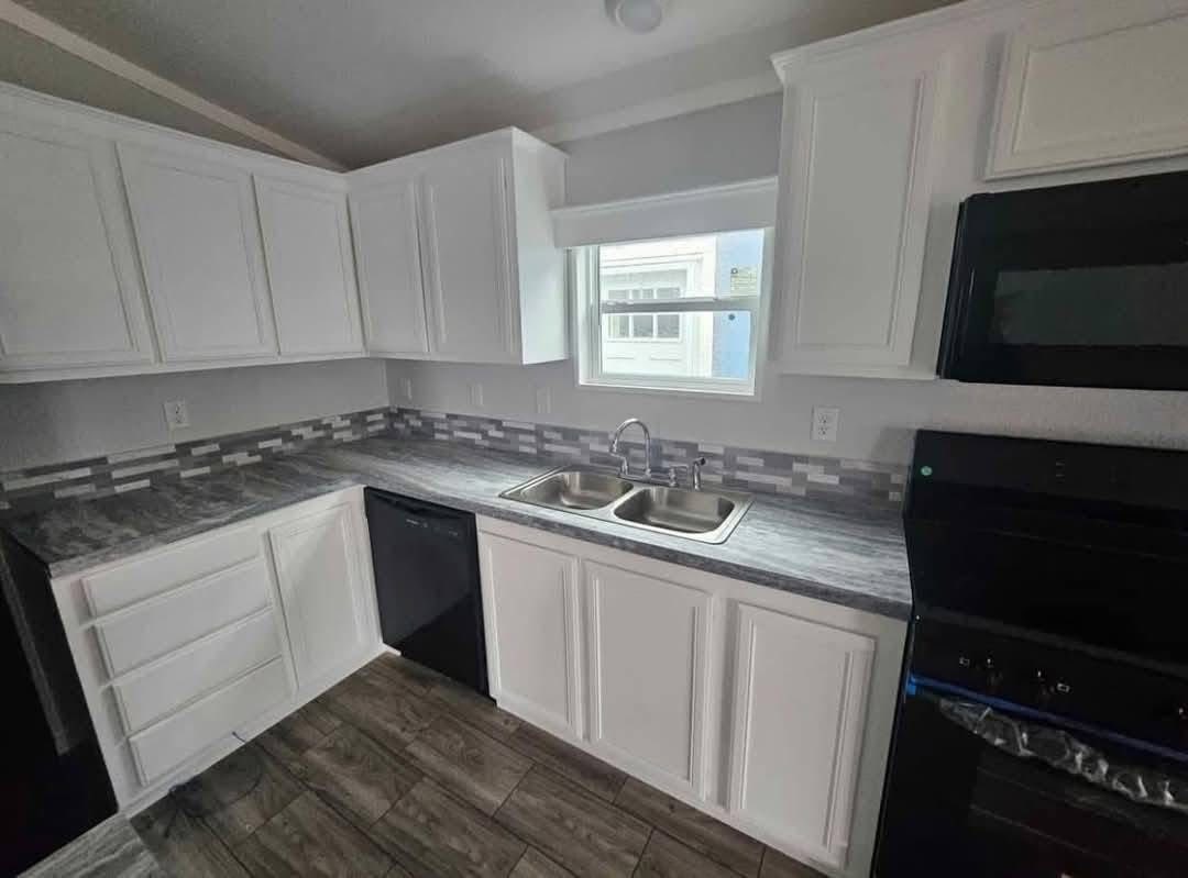 Modern kitchen with white cabinets, gray marble countertops, and a dual sink. A small window provides natural light, and a black stove and dishwasher complete the space.