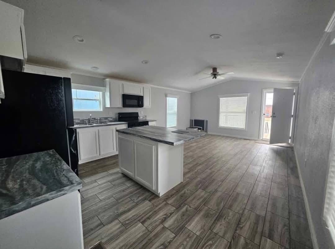 Spacious kitchen and living area with white cabinets, granite countertops, wood-like tile floor, ceiling fan, and natural light through large windows.