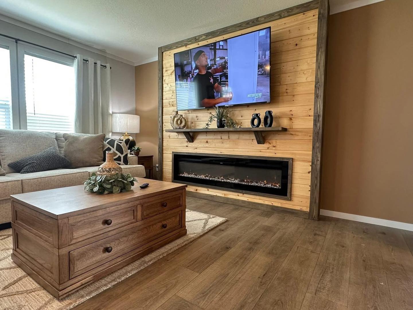 Cozy living room with a wall-mounted TV above a wooden paneled electric fireplace. A beige sofa, wooden coffee table, and decor add warmth and charm.