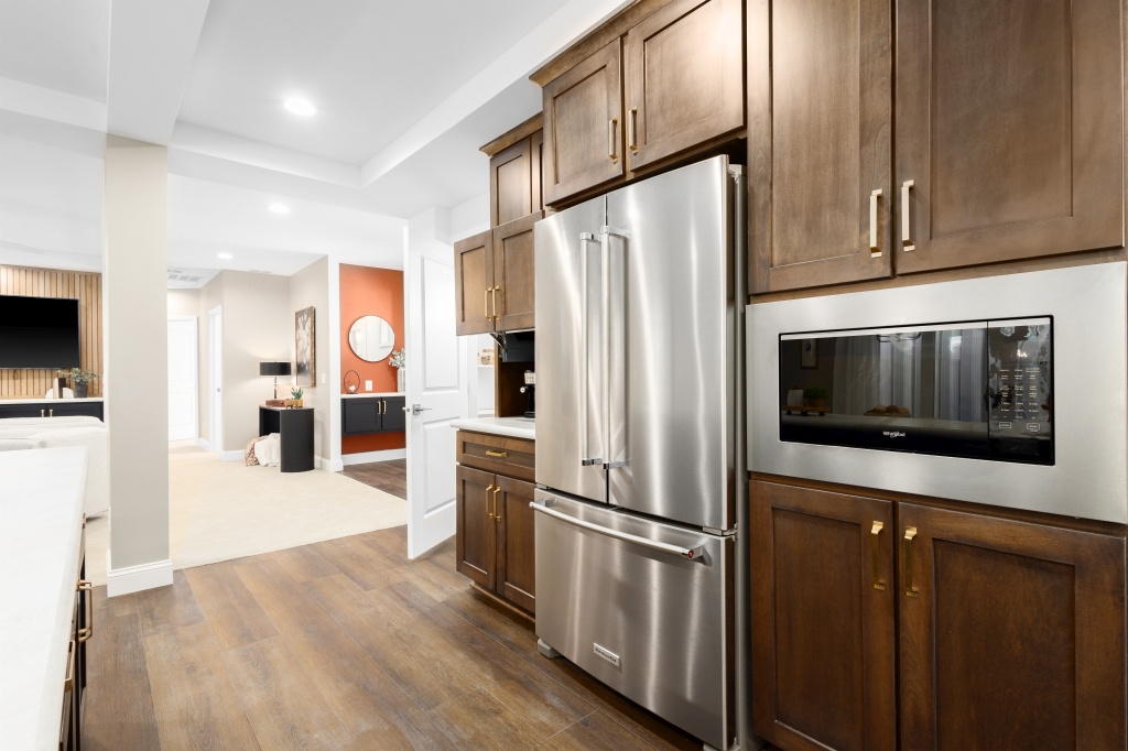Modern kitchen with stainless steel fridge and built-in microwave, surrounded by dark wood cabinets. Leads to an open living area with warm tones.