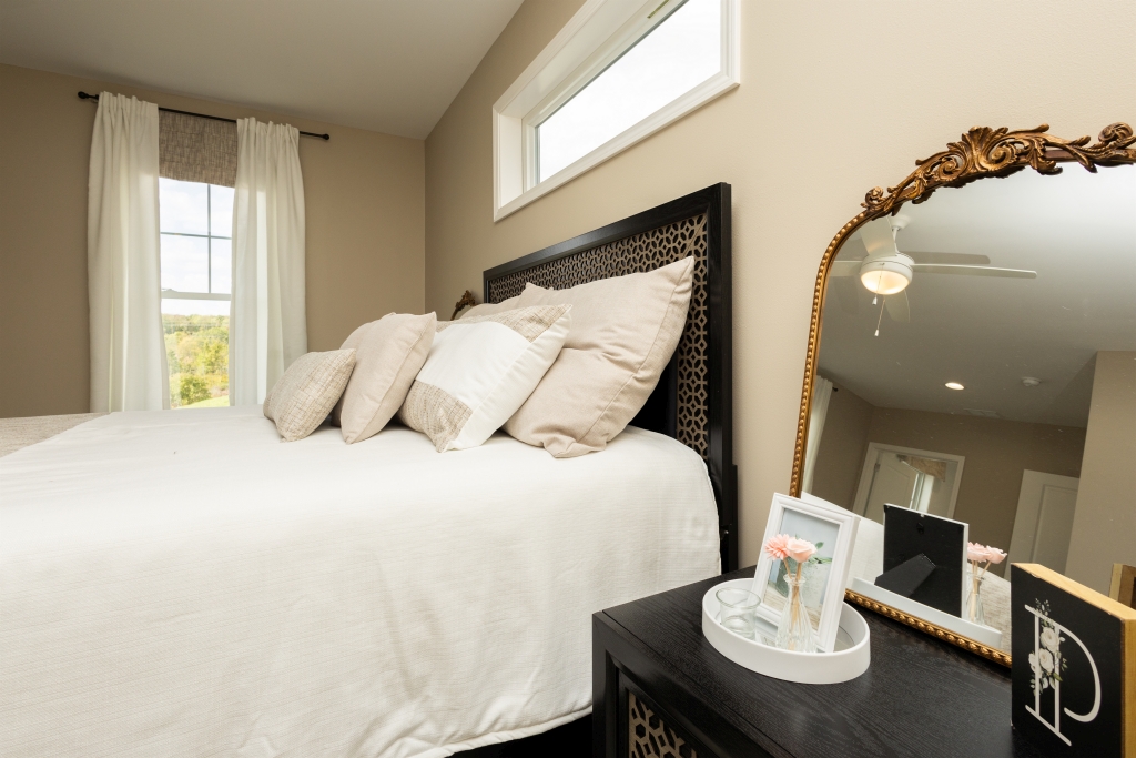 Cozy bedroom with a bed adorned with neutral pillows and a white blanket. A gold-edged mirror reflects a ceiling fan. Soft natural light enhances the calm ambiance.