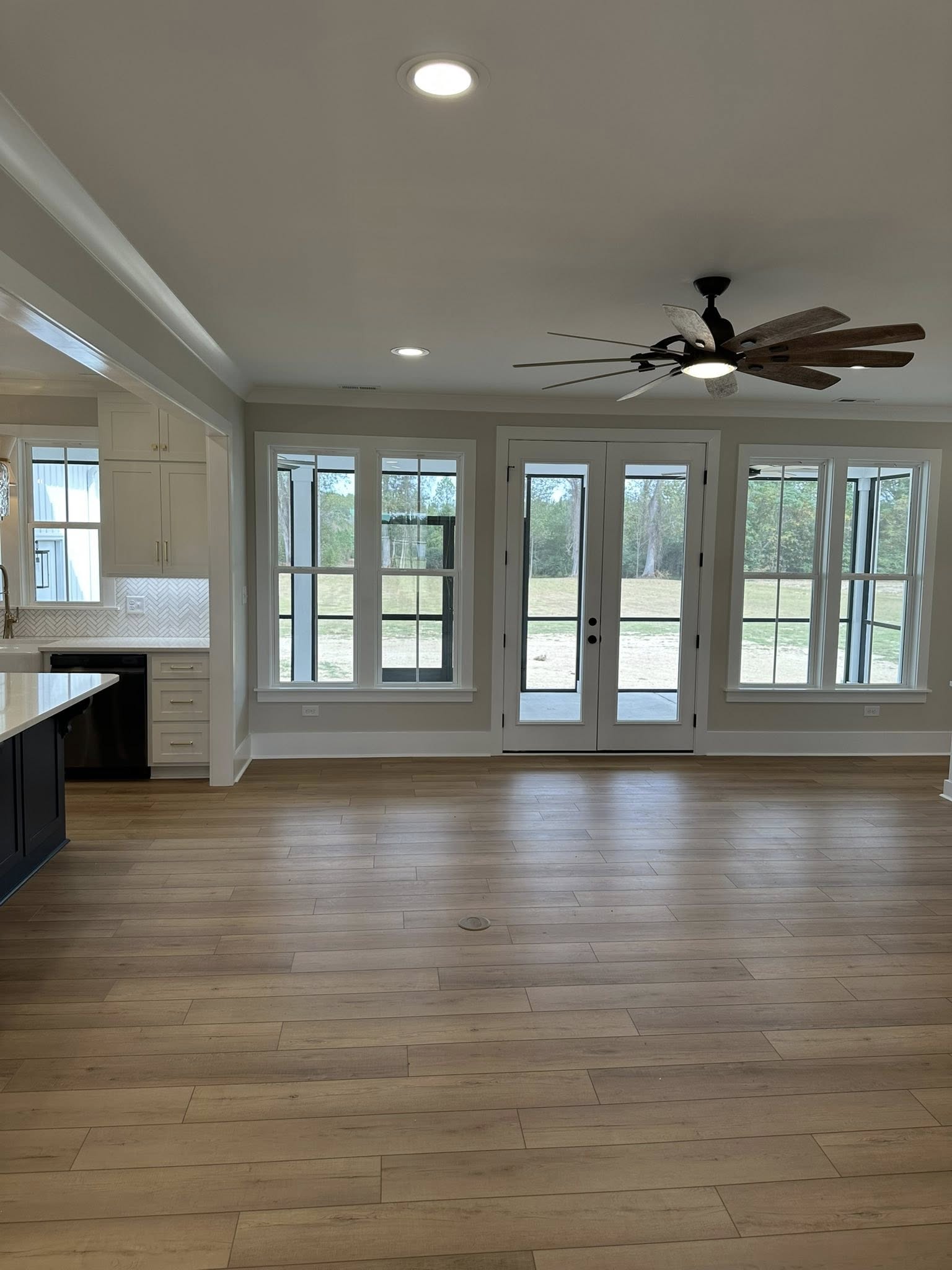 Spacious living area with light wood floors, large windows, and a glass door overlooking greenery. Ceiling fan and a glimpse of kitchen cabinets. Calm atmosphere.