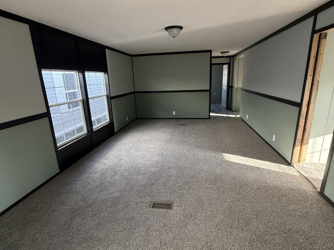 Empty room with gray walls, black trim, and two large windows. Beige carpet and a simple ceiling light give a calm, clean atmosphere.
