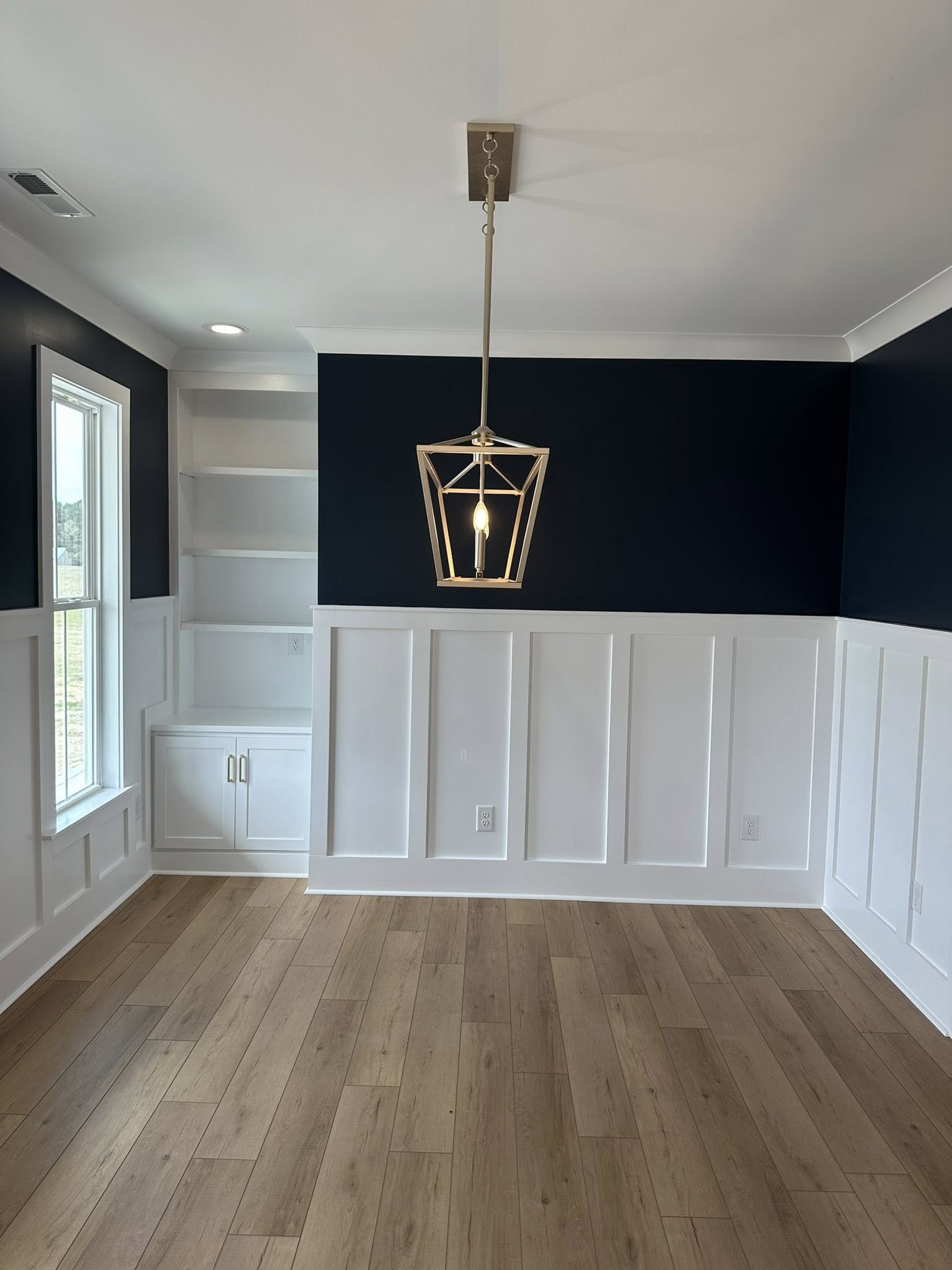 A modern room features a wooden floor, navy and white paneled walls, a geometric pendant light, and built-in shelves by a large window, creating an elegant ambiance.