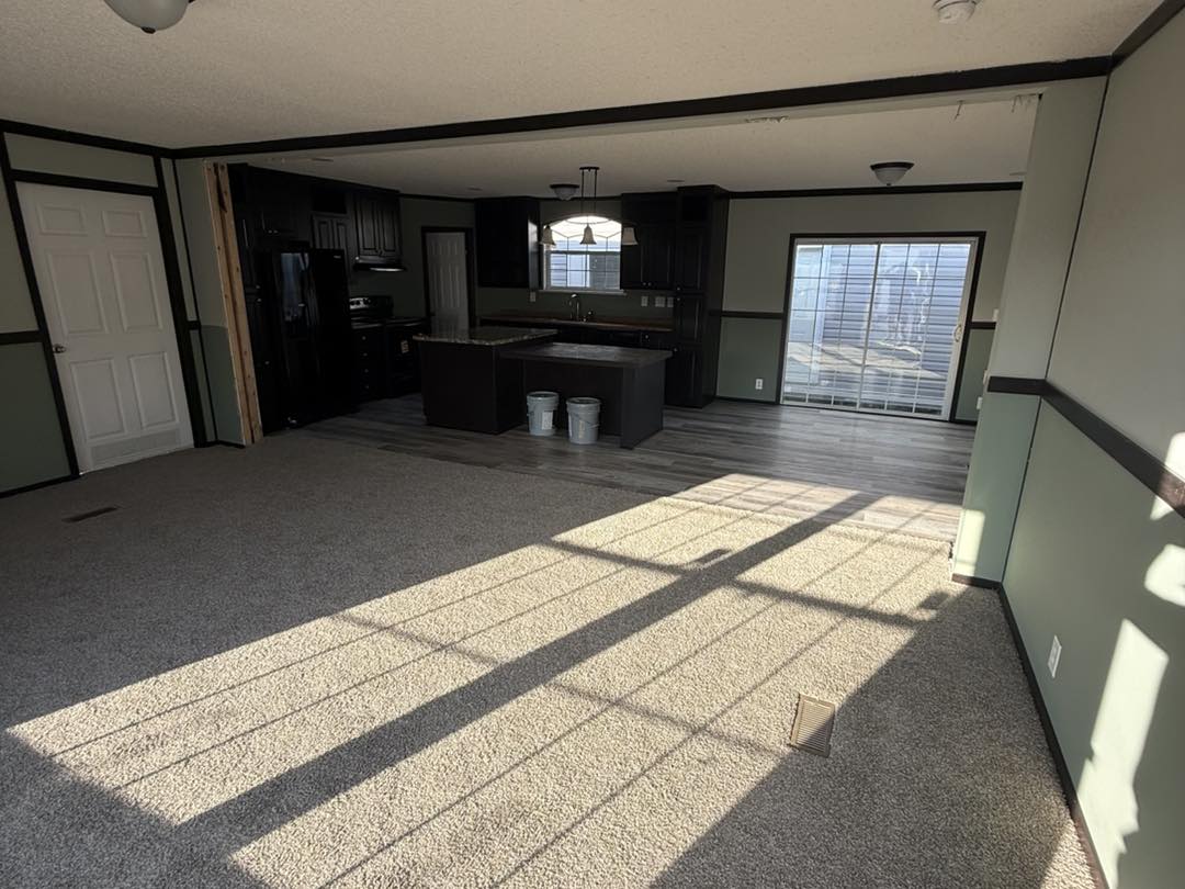 Spacious living room with beige carpet and sunlight streaming through a large window. In the background, a modern kitchen with dark cabinets.
