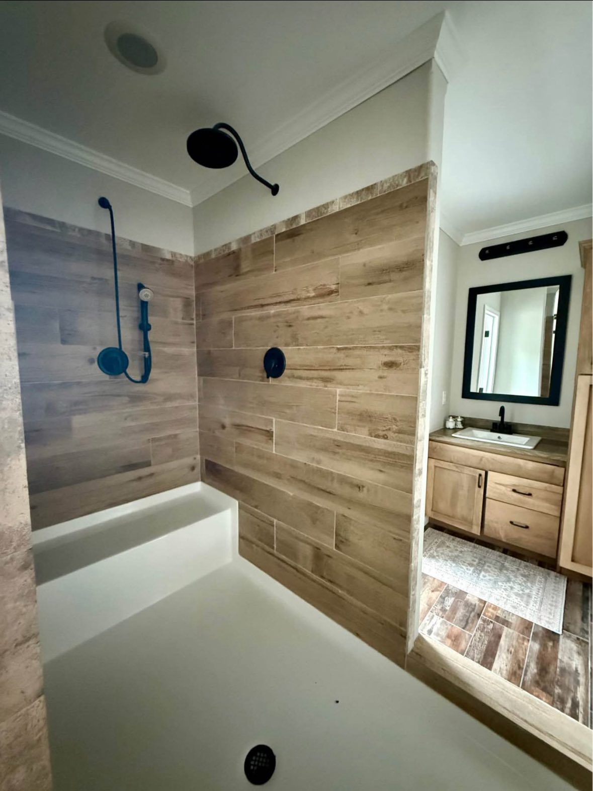 Modern bathroom with a large walk-in shower featuring wood-patterned tiles. A sink with a mirror is visible in the adjoining room, creating a cozy ambiance.