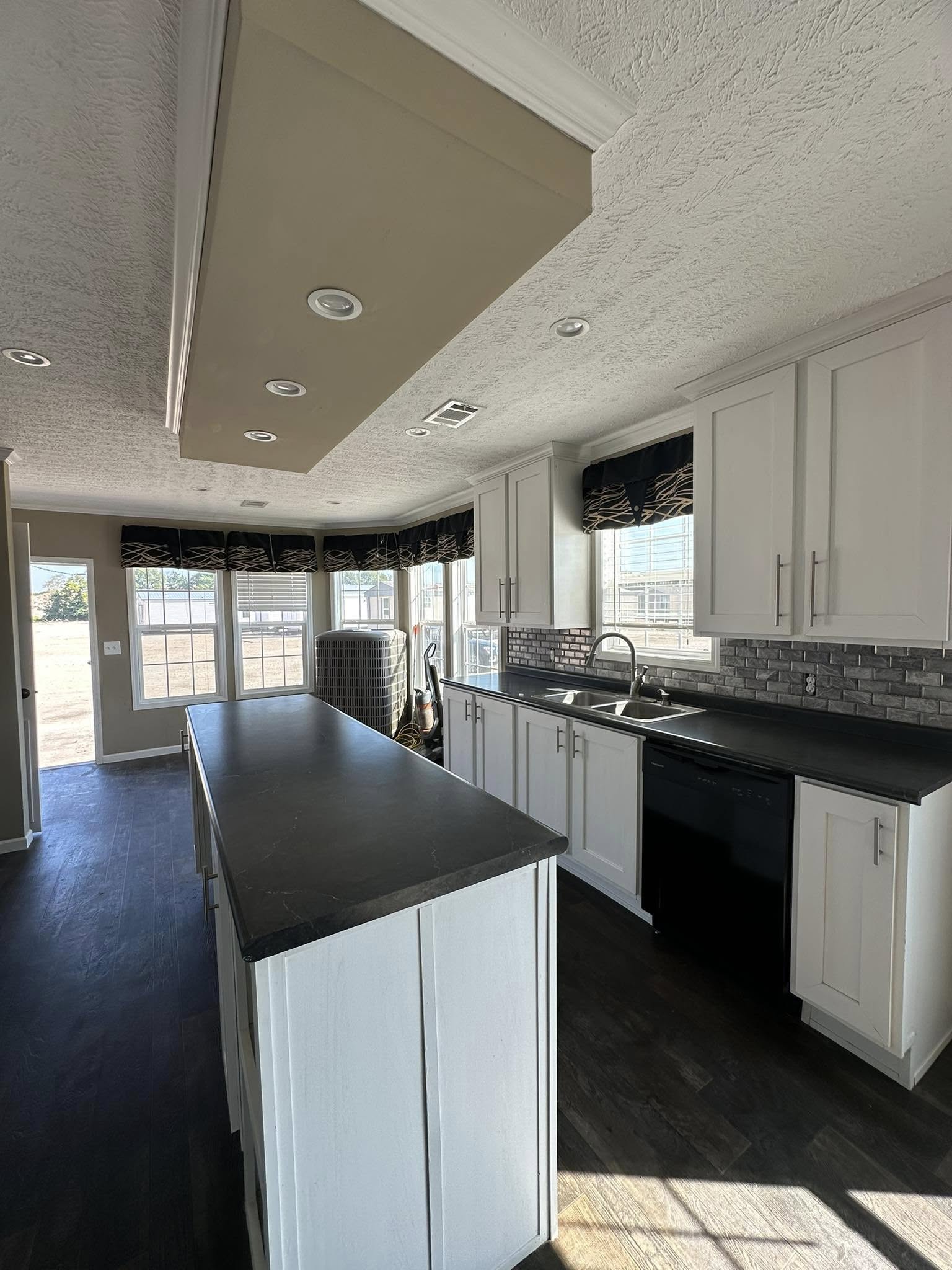 Modern kitchen with white cabinetry, dark countertops, and a black tile backsplash. Large windows with patterned valances bring in natural light.