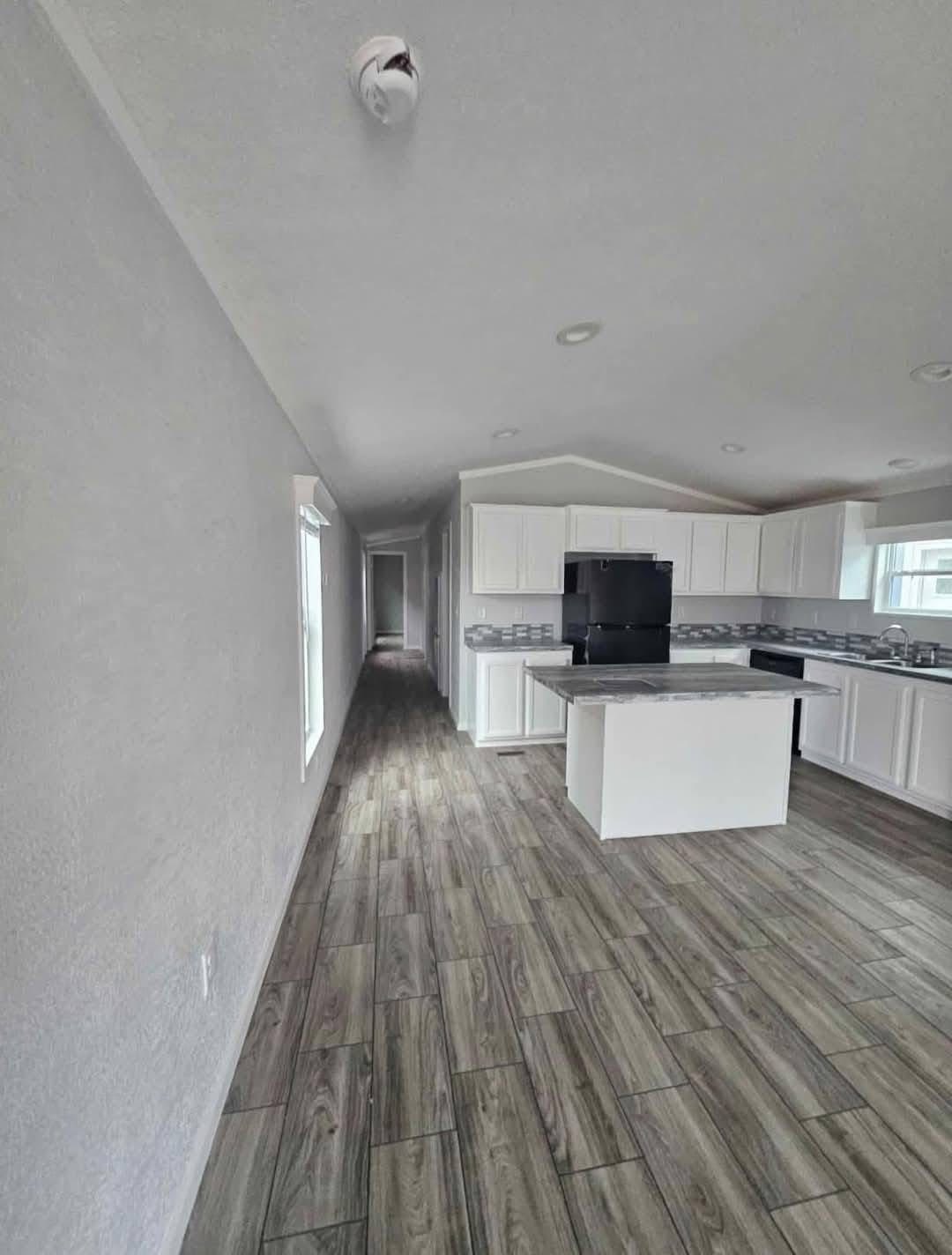 Narrow kitchen with white cabinets, black appliances, and island. Wood-patterned floor, recessed lighting, and a bright hallway lead to the back. Cozy and modern.
