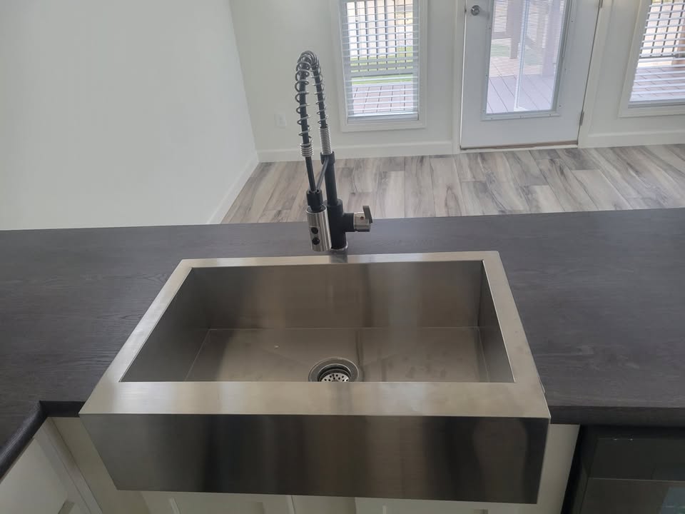 Stainless steel farmhouse sink with a coiled spring faucet on a dark countertop. Background shows light wood flooring and glass doors. Modern kitchen feel.