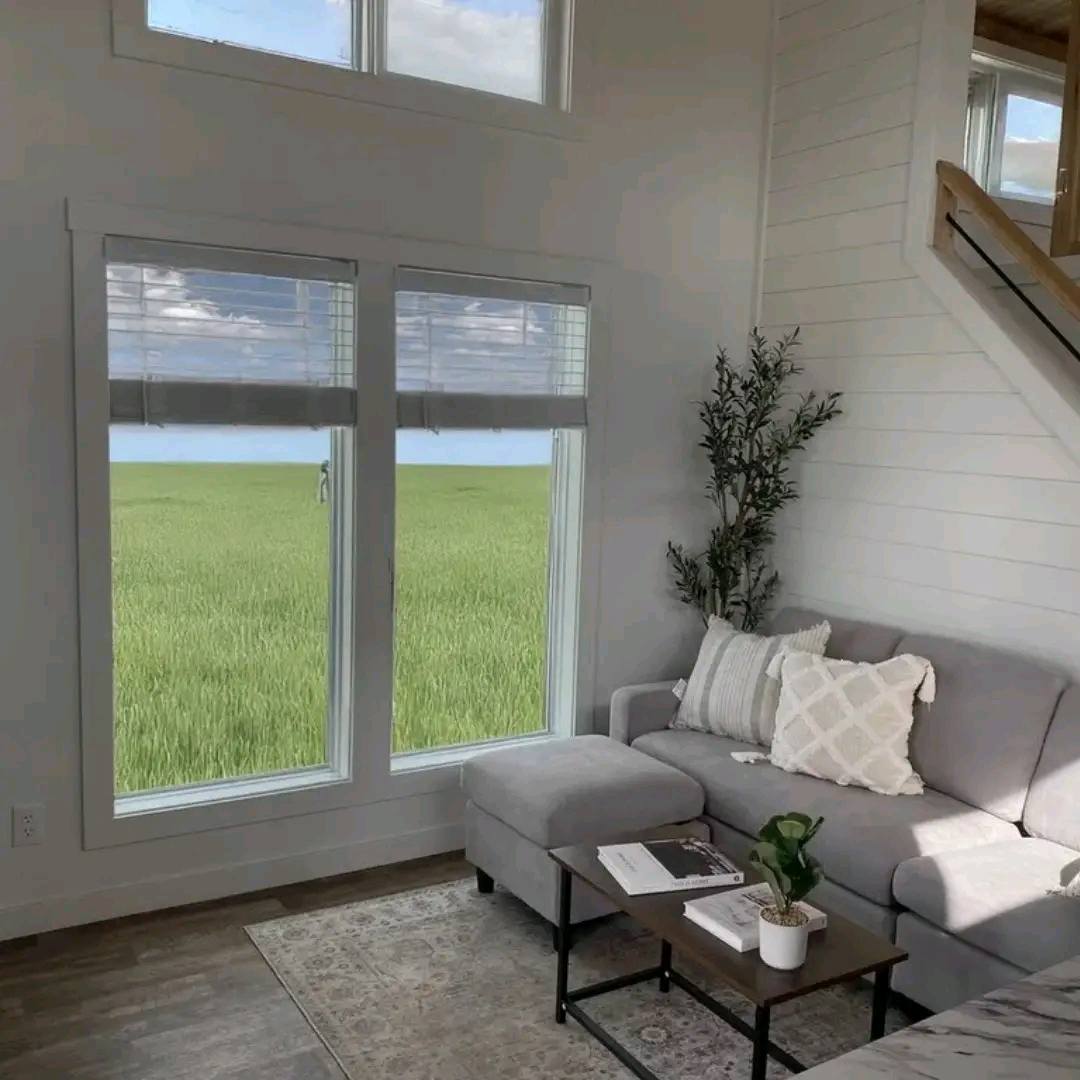 Bright, cozy living room with gray sectional sofa, decorative pillows, a small table with a plant, and large windows showcasing green fields and blue sky.