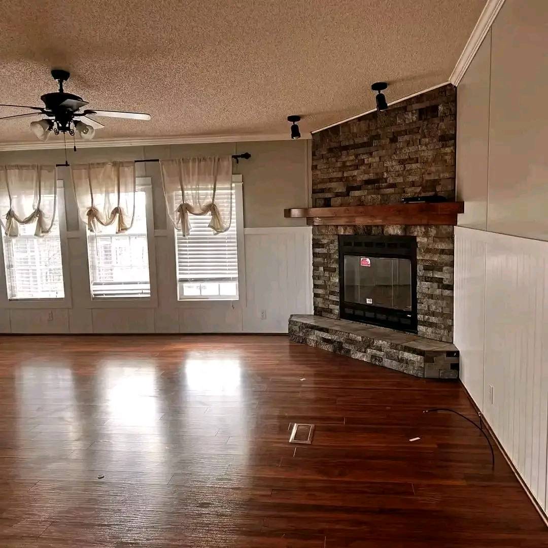 Spacious living room with polished hardwood floors, a brick fireplace with a wooden mantel, and sheer curtains over large windows. Cozy and inviting ambiance.