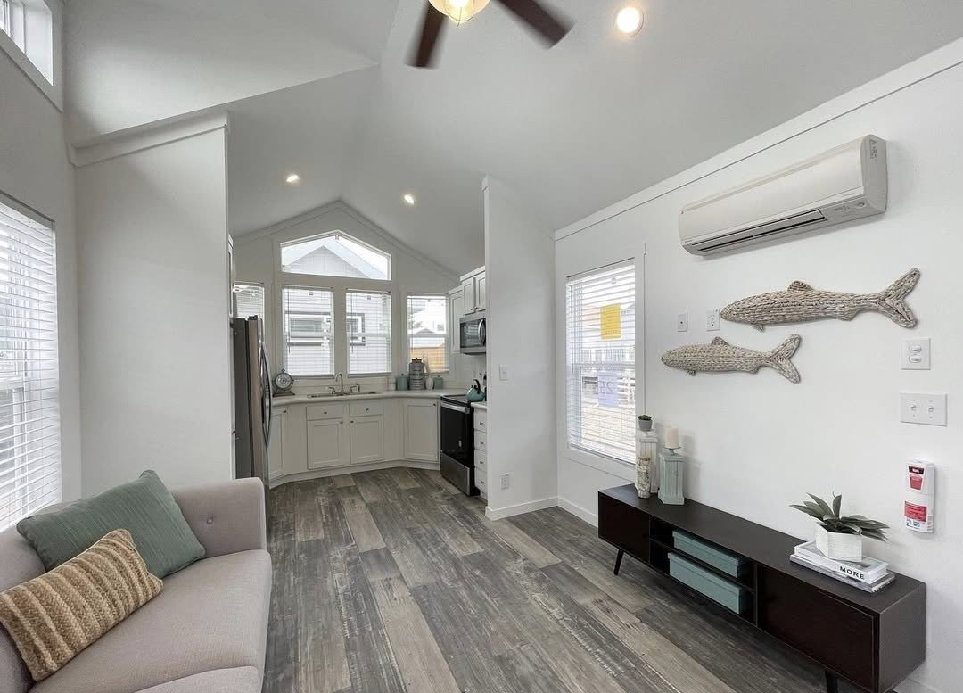 Bright, modern living room with vaulted ceiling and large windows. Features a small kitchen, cozy sofa, wood floors, and wall decor shaped like fish.